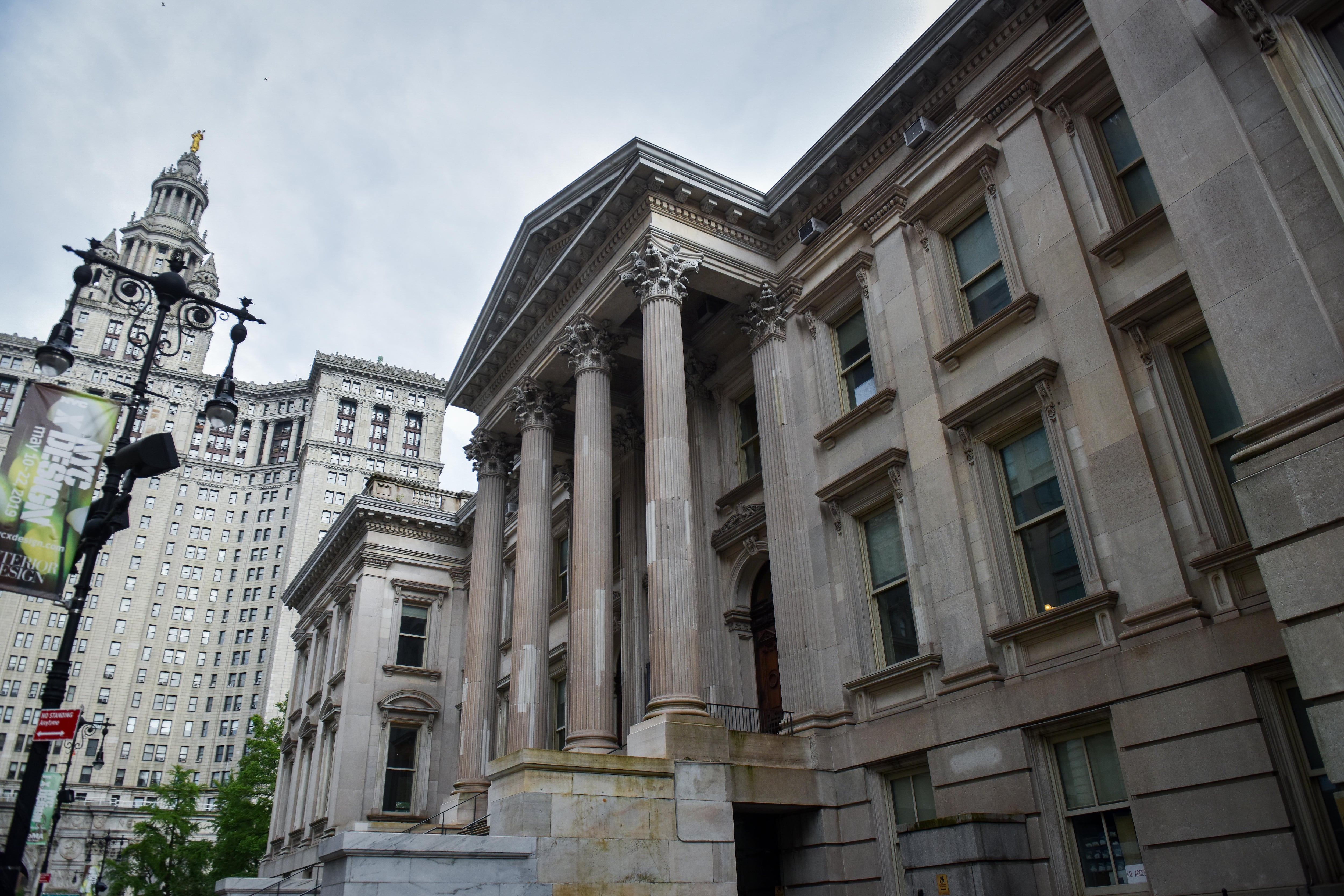 A large, white stone building with columns with other skyscrapers in the background.