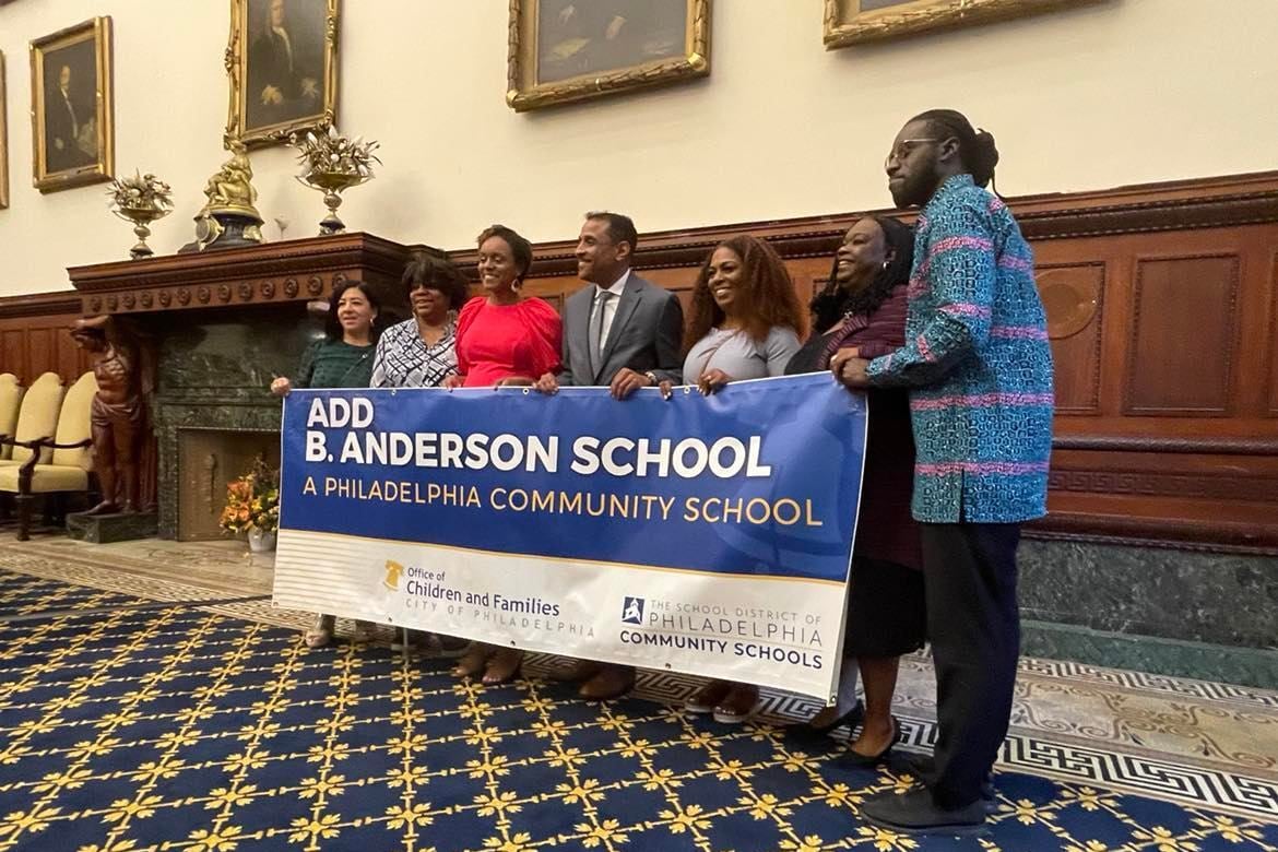 Seven people hold up a blue and white banner with the name “Add B. Anderson School” written in white.