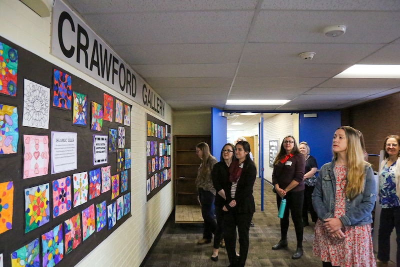 A group of people stand in a large room looking at a decorated wall on the left side of the frame.