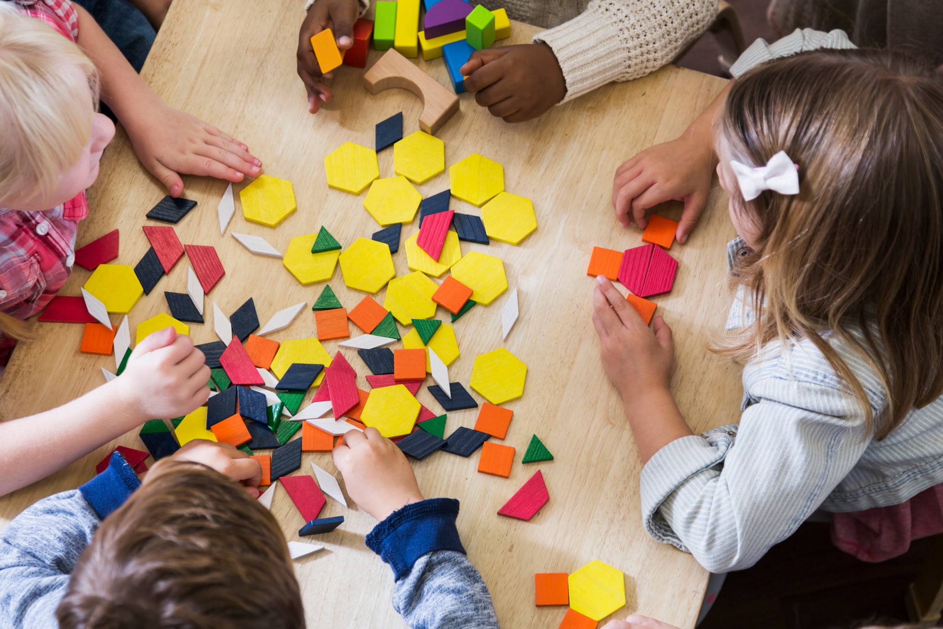 An overhead view of a group of five preschoolers sitting at a table playing with colorful blocks and geometric shapes. They are unrecognizable since we see the tops of their heads and their hands. They are in a preschool or kindergarden classroom.