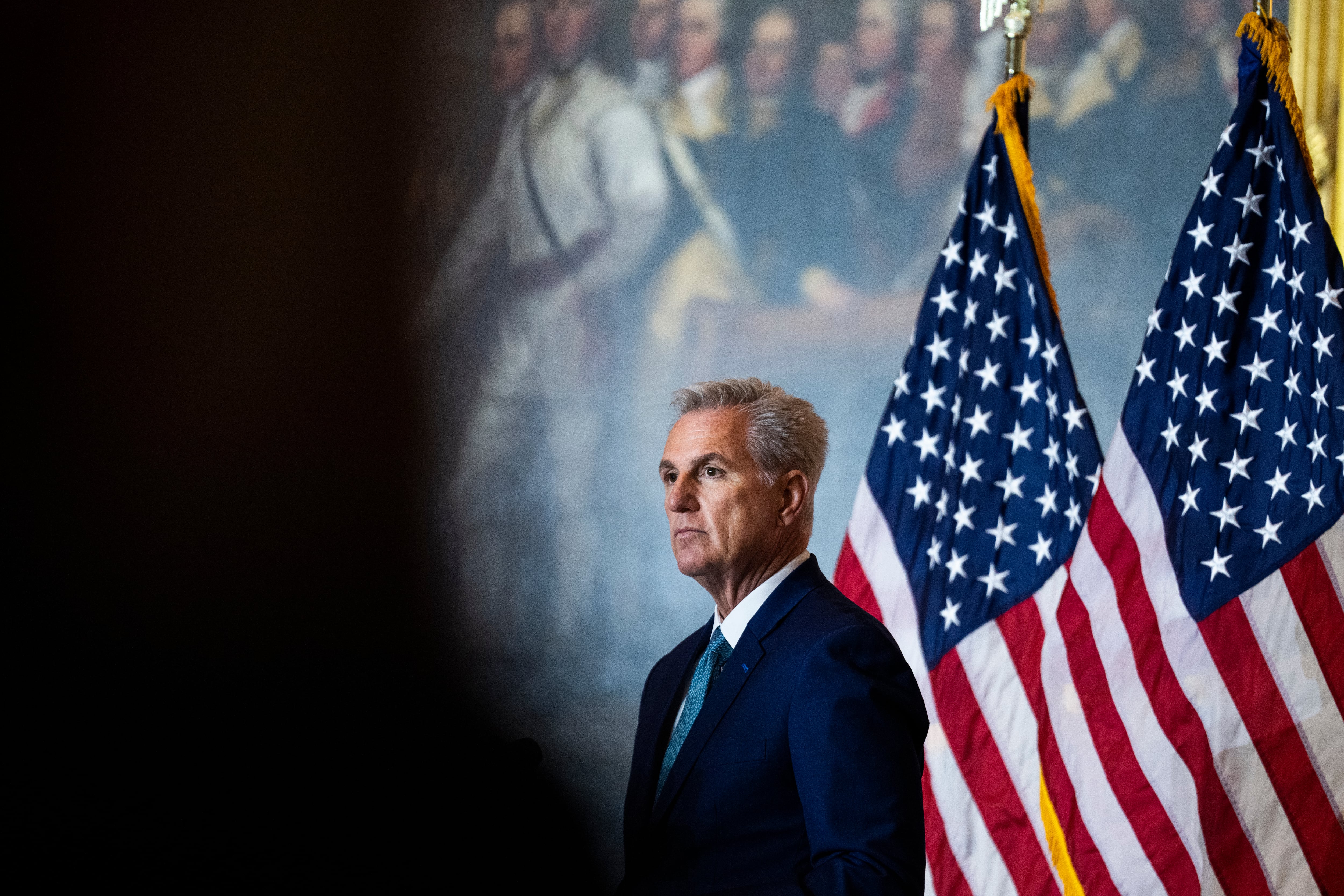 A man in a suit stands in front of two American flags