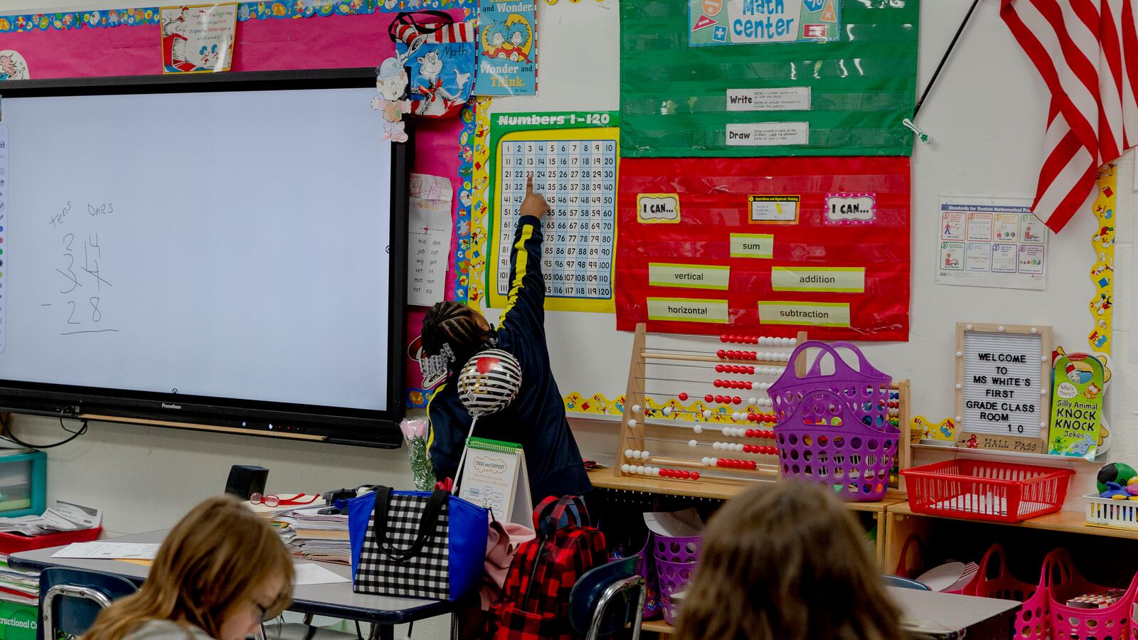 A young girl points at a number chart next to a smart board at the front of a classroom.