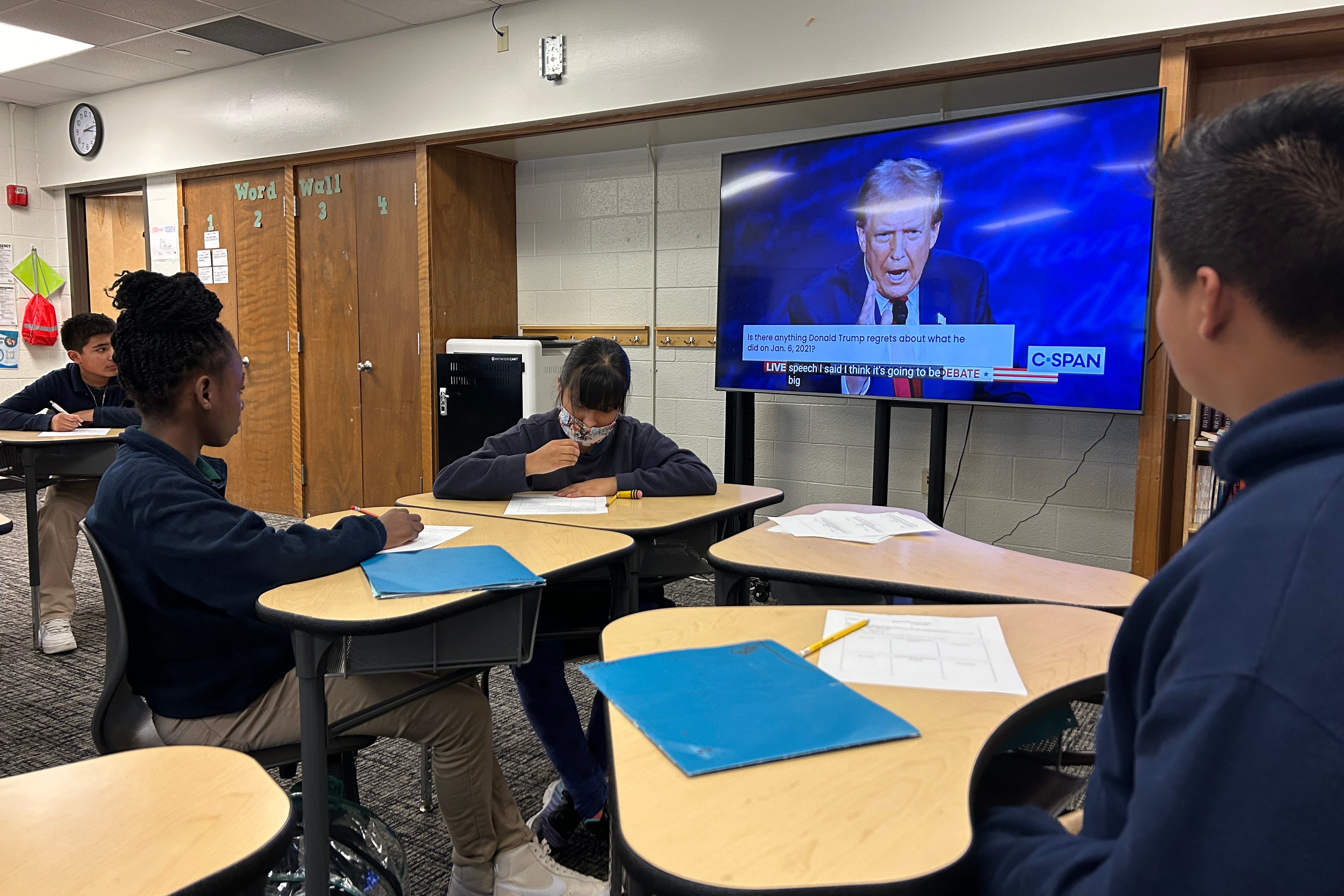 Three students sit at desks in a classroom while a TV shows a picture of Donald Trump in the background.