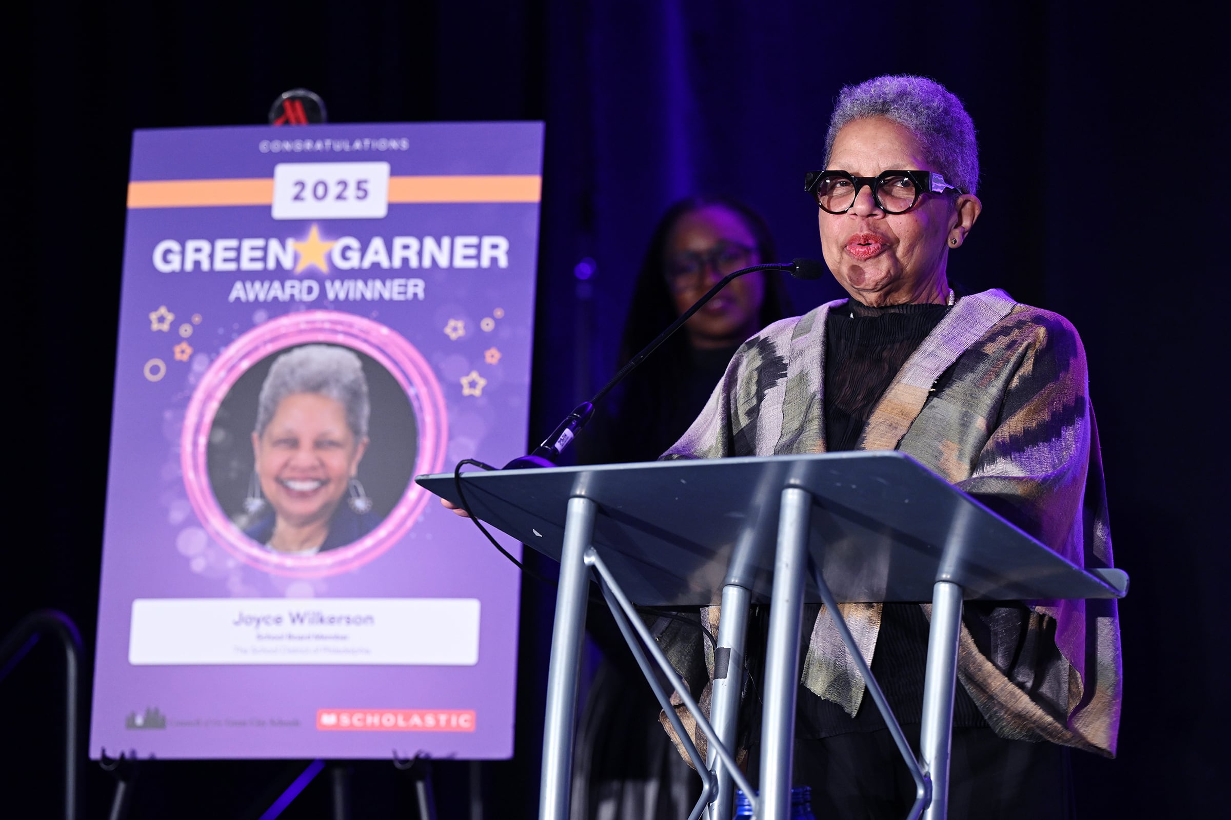 A photograph of a Black woman with short hair and wearing glasses stands proudly on stage speaking into a microphone with a large poster to the left and a person in the background.