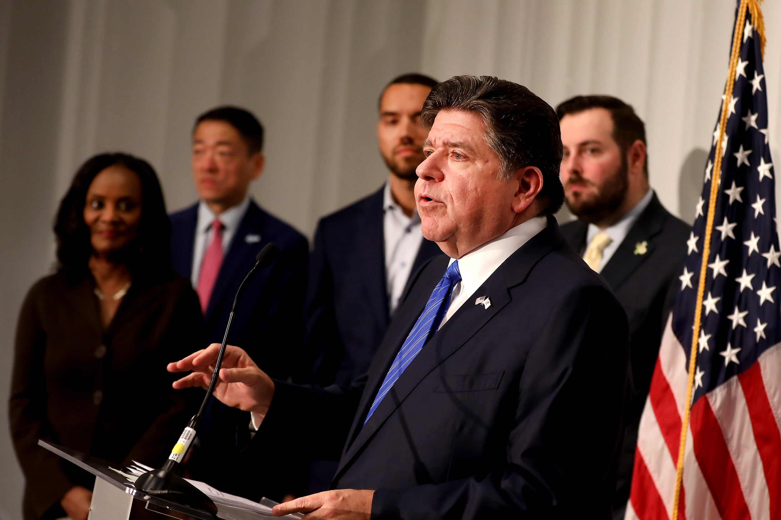 A man in a suit speaks from a podium with a group of people stands in the background and next to an American flag.