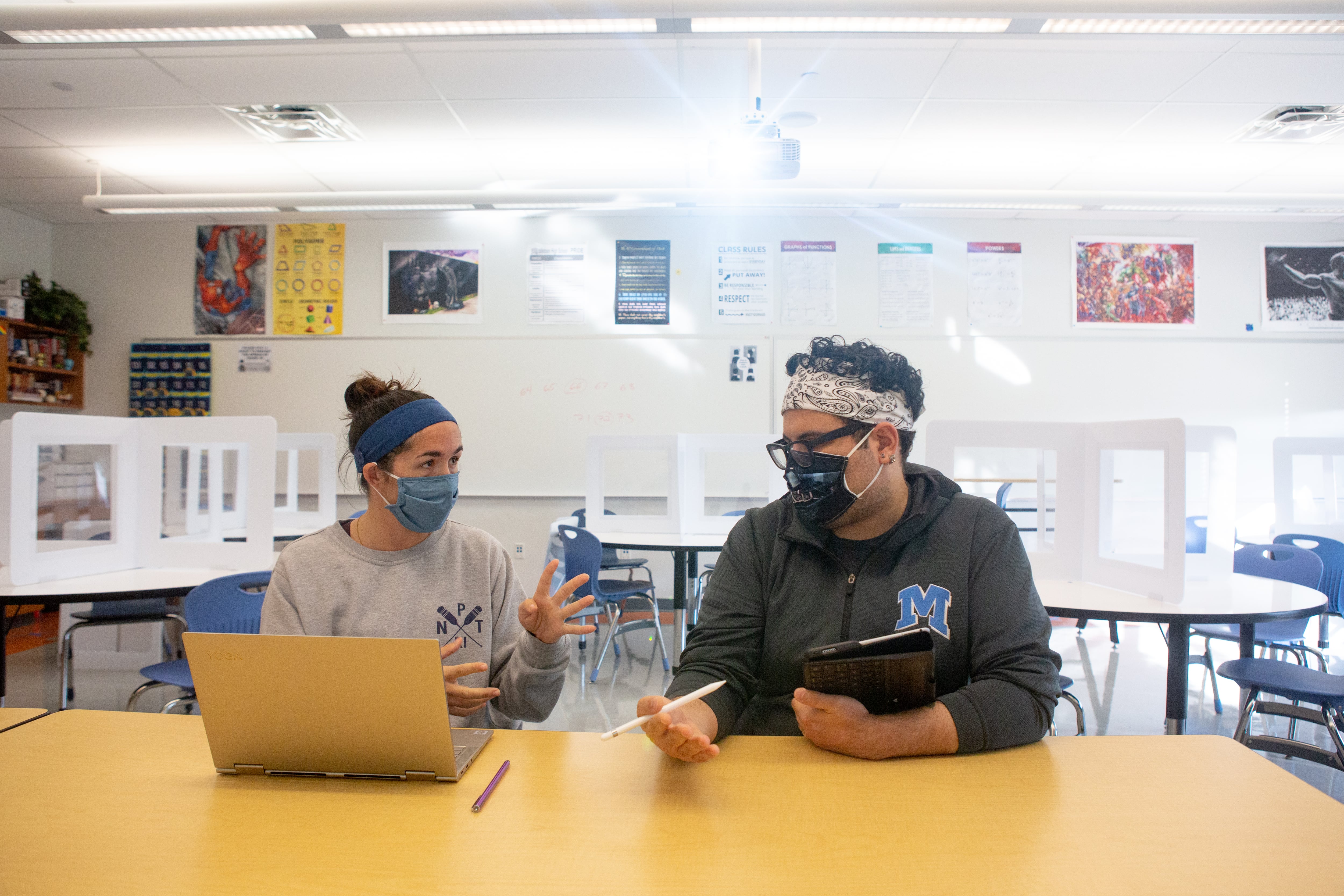 Two math teachers plan upcoming lessons. They are sitting at a table behind a laptop. Both are wearing masks.