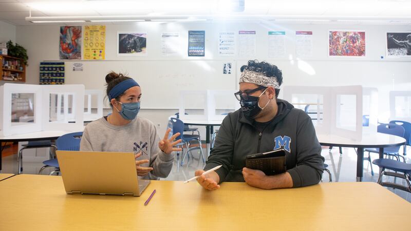 Two math teachers plan upcoming lessons. They are sitting at a table behind a laptop. Both are wearing masks.