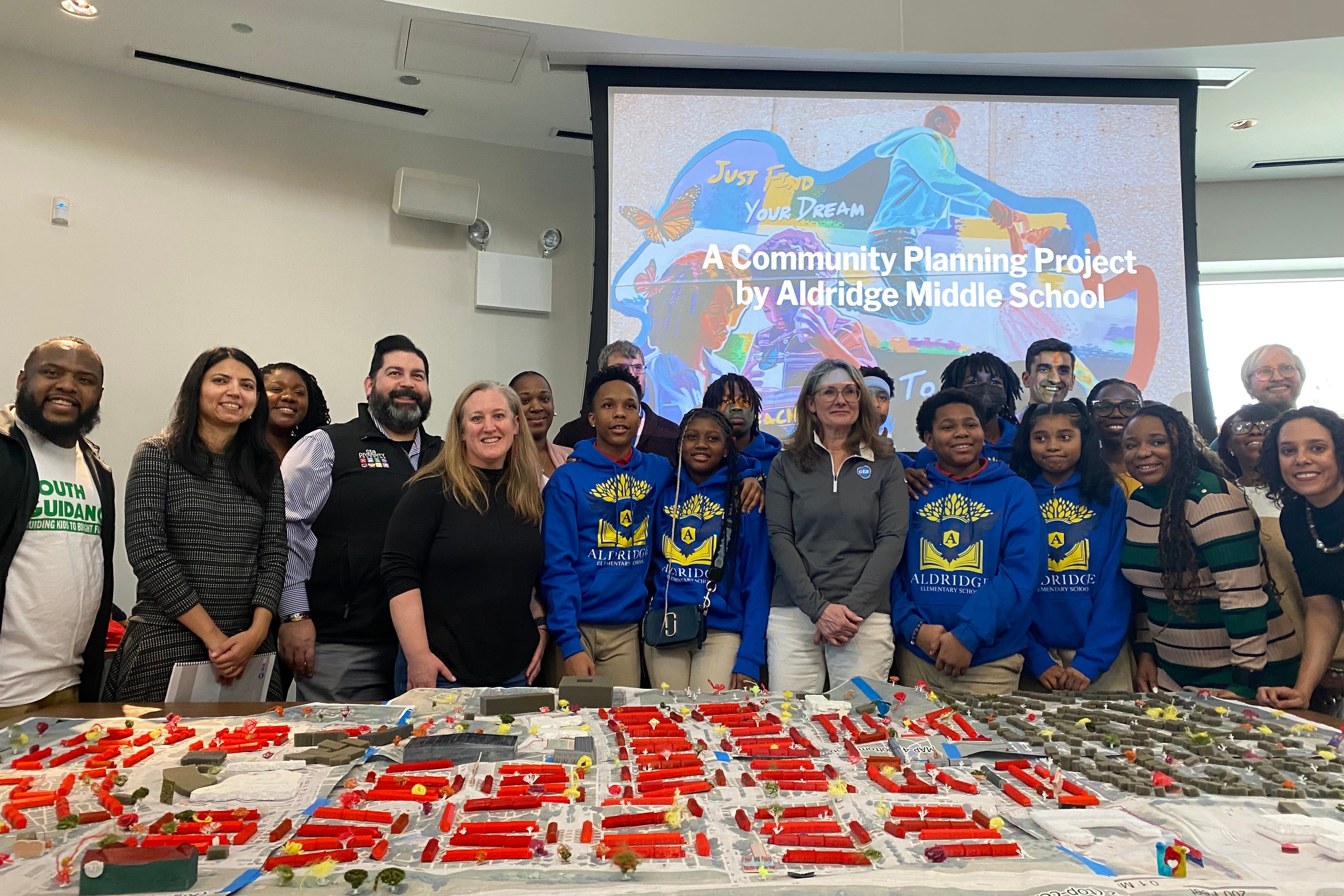 A group of students and adults pose for a photograph behind a table of 3D models with a screen in the background.