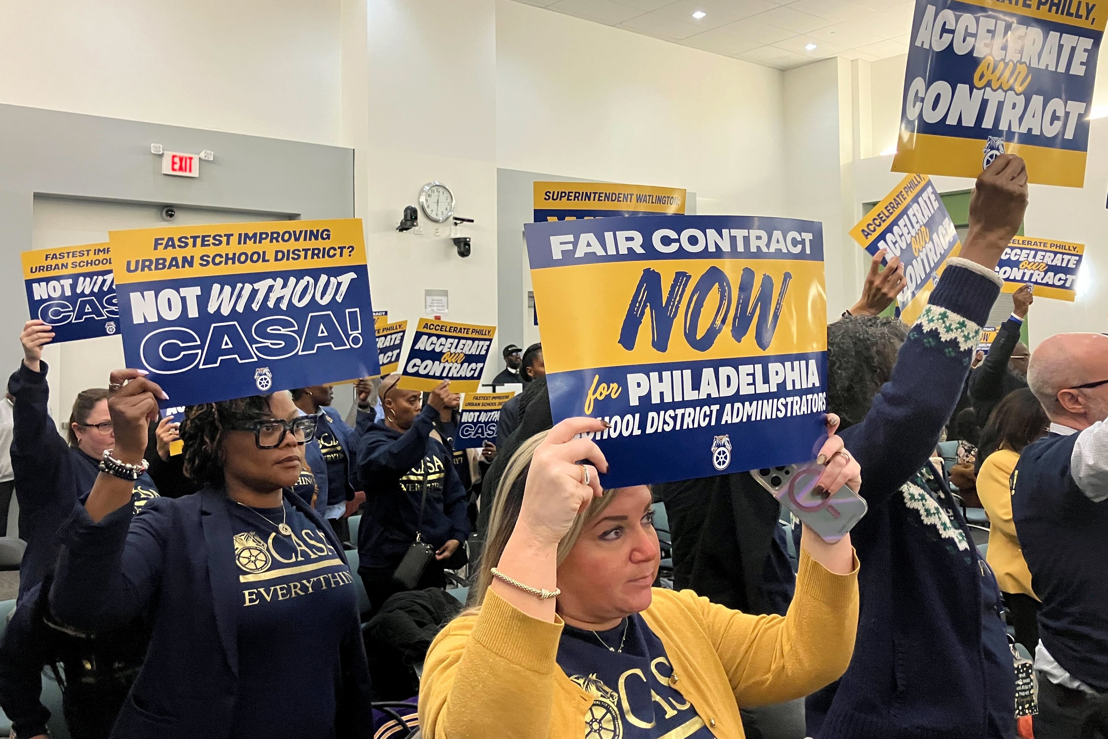A photograph of a meeting room full of adults, some of them are holding up protest signs.