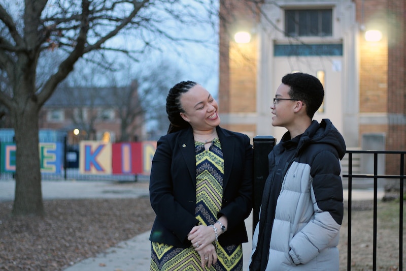 An adult woman stands next to a middle school boy outside and pose for a portrait in front of a school.