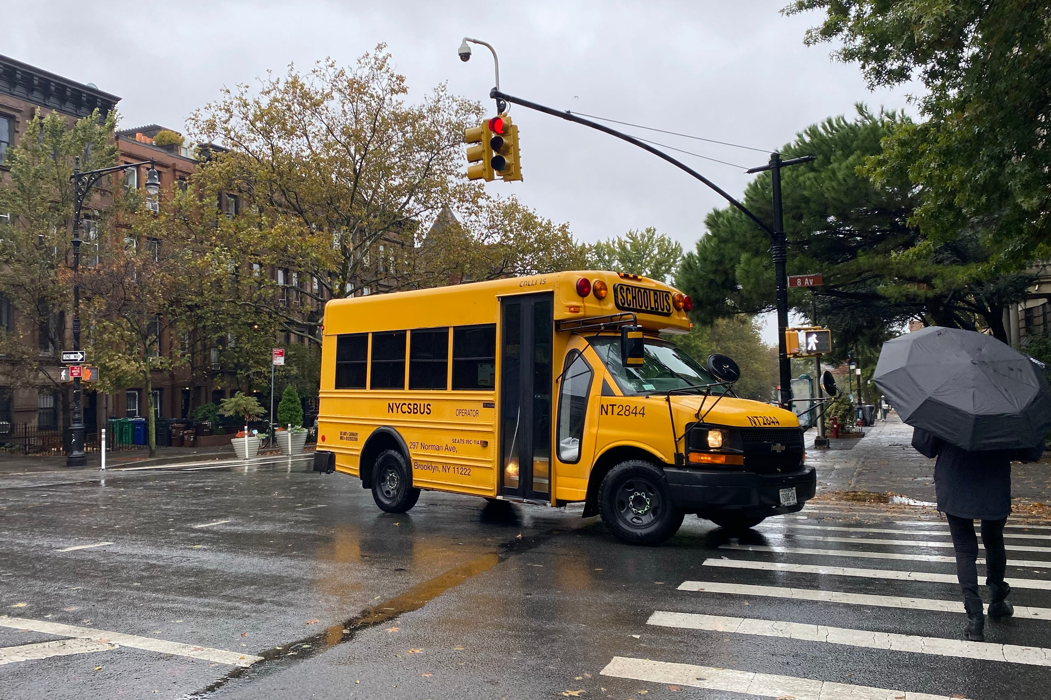 A photograph of a small yellow school bus crossing an intersection while a person holding an umbrella crosses at the cross walk with grey sky above.