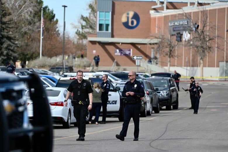 Several police officers in uniform walk through a parking lot in front of a high school building, which looms in the background.