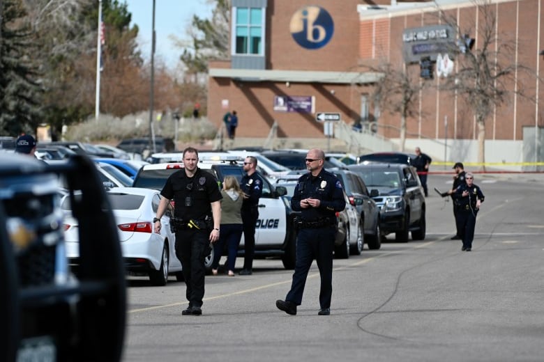 Several police officers in uniform walk through a parking lot in front of a high school building, which looms in the background.