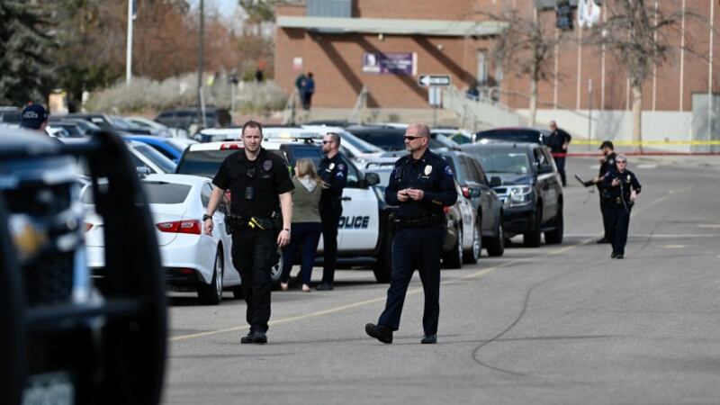 Several police officers in uniform walk through a parking lot in front of a high school building, which looms in the background.