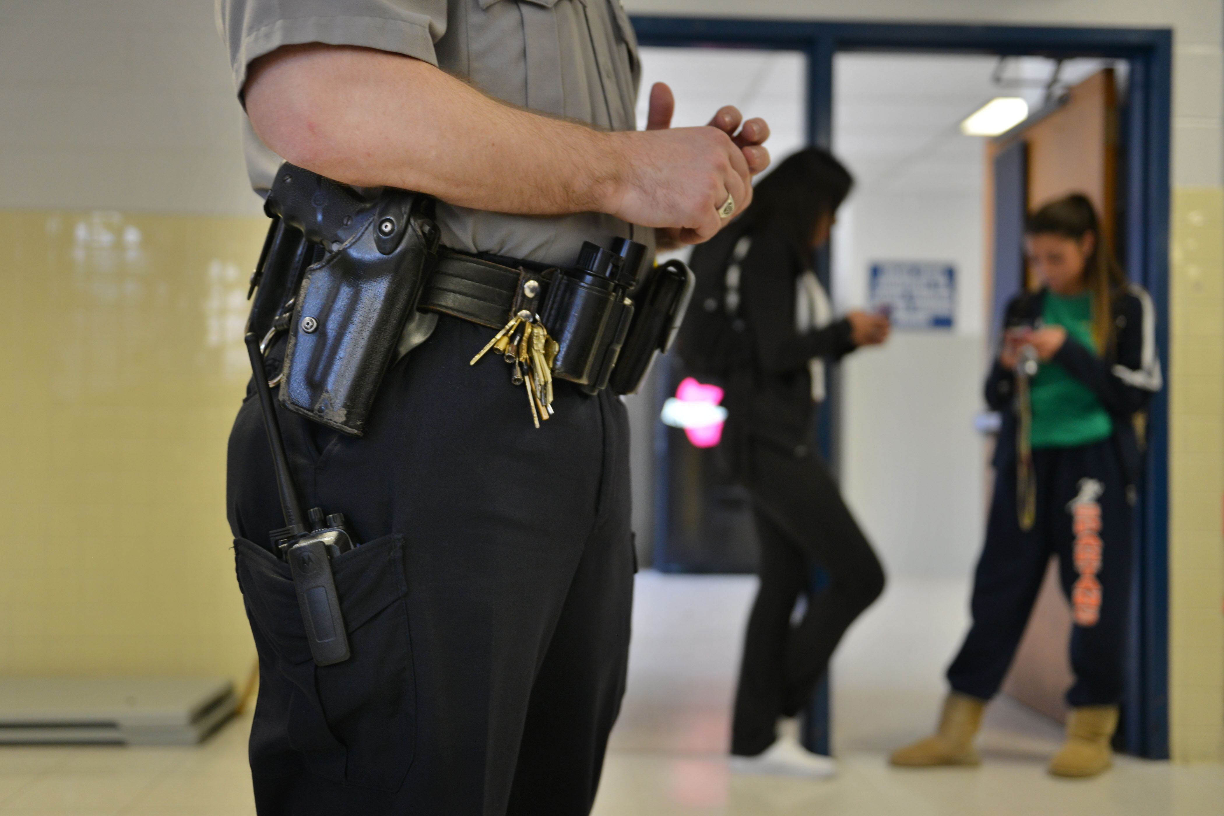 A close up of a school police officer's utility belt in a school hallway with two students in the background.