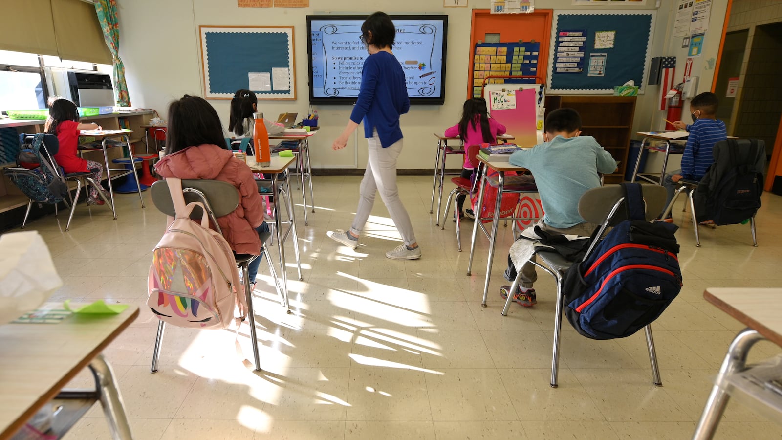 Students sit at their desks in a classroom during in-person instruction as their teacher walks through the room. There is a ray of light reflecting off of the floor of the classroom in between two rows of desks.