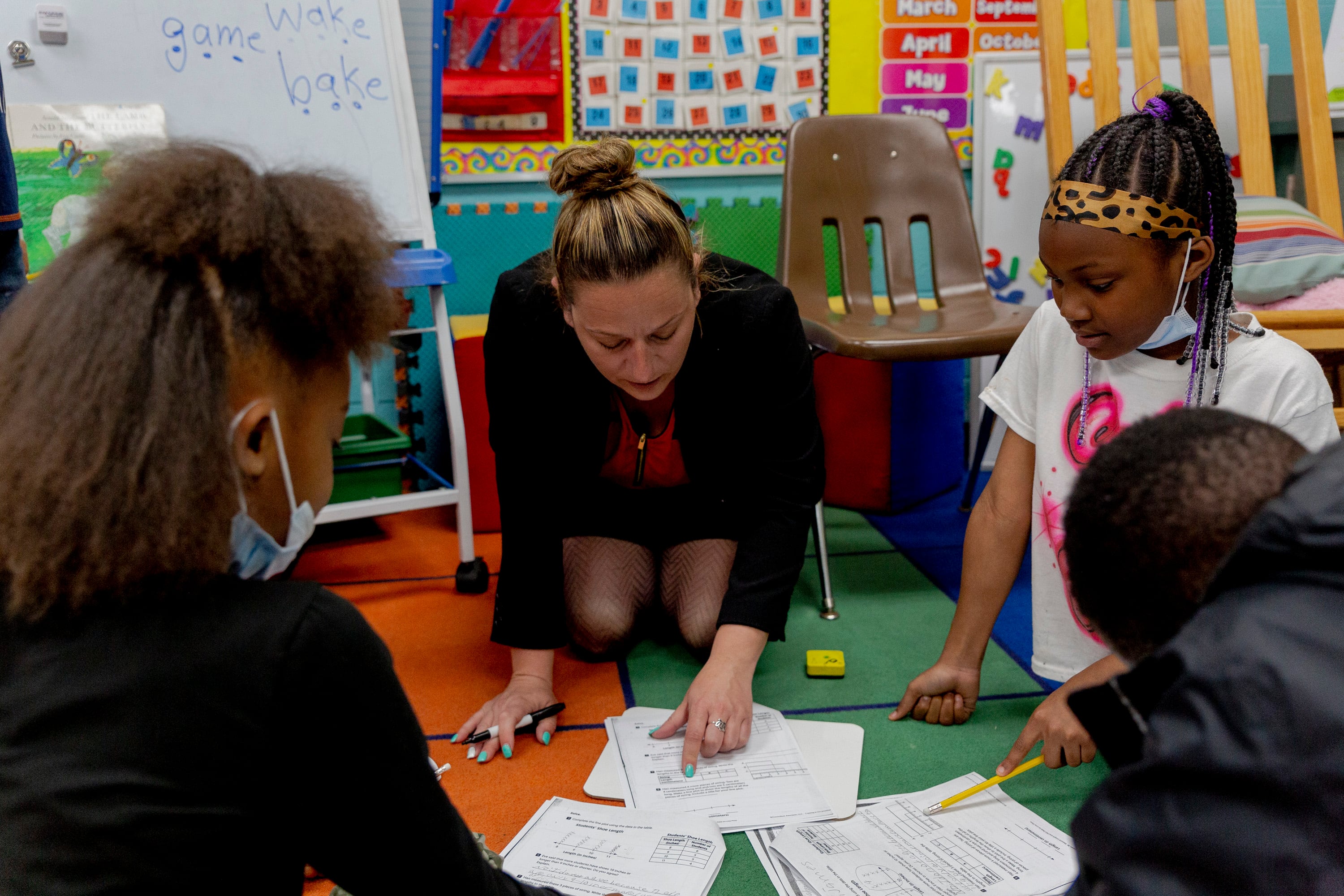 Woman wearing suit and kneeling on a colorful carpet in a classroom points to paper worksheet as a girl wearing leopard print headband looks on