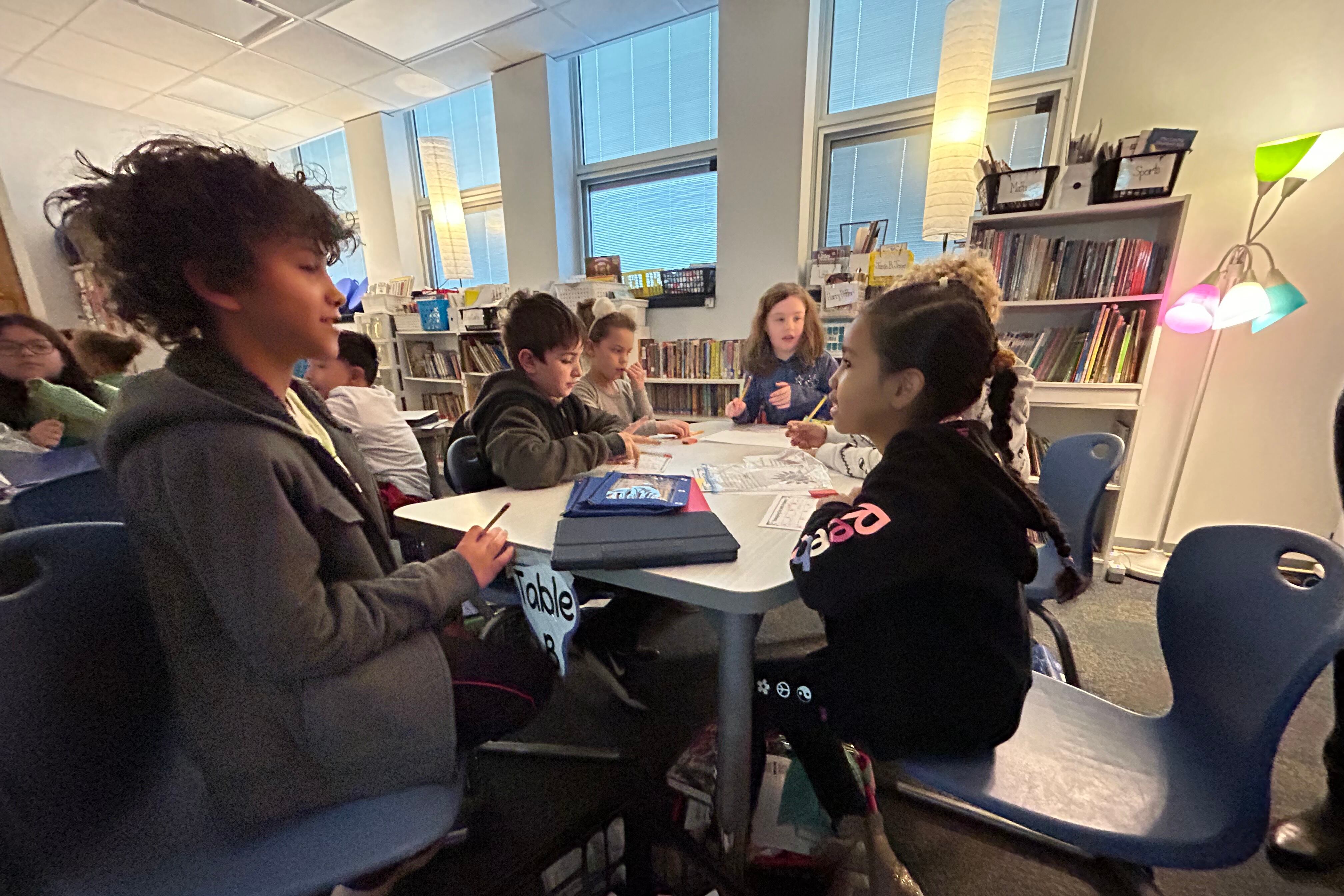 Six elementary students sit talking at a table with papers and pencils.