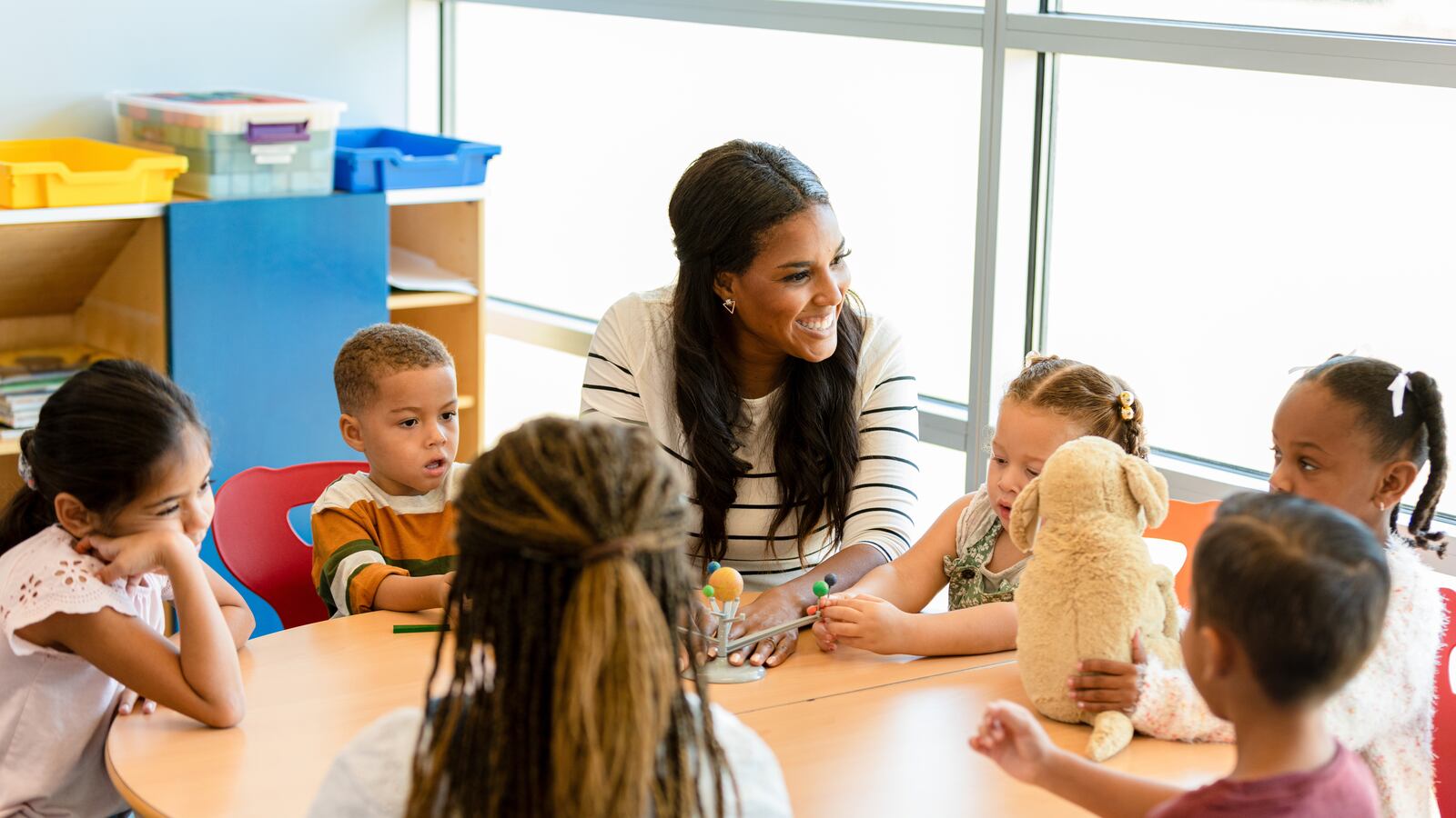 A group of young children sit around a round table as their teacher teaches them using a model of the solar system.