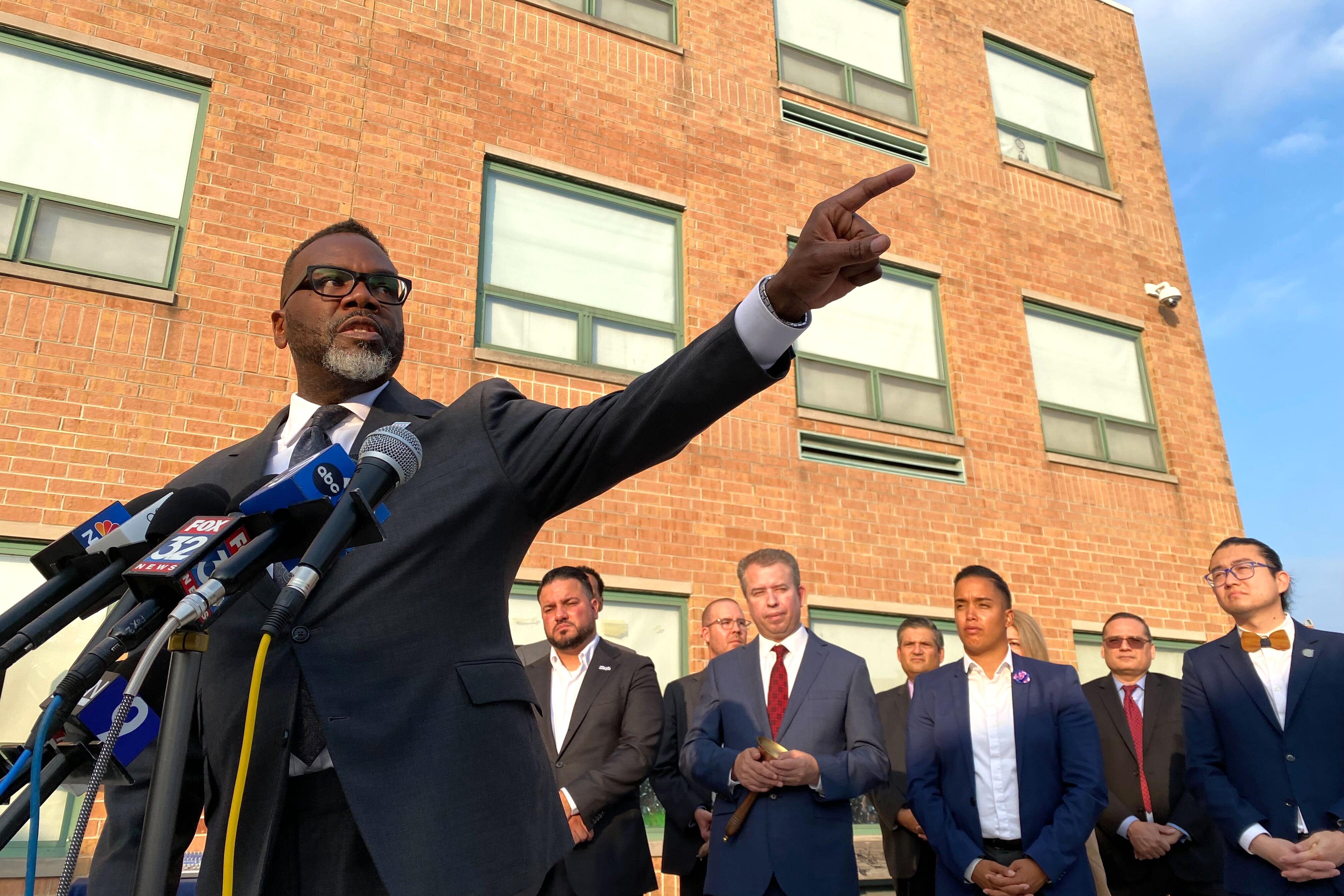 A man in a dark suit speaks from behind a podium and points to the side while a line of people in suits stand in the background.