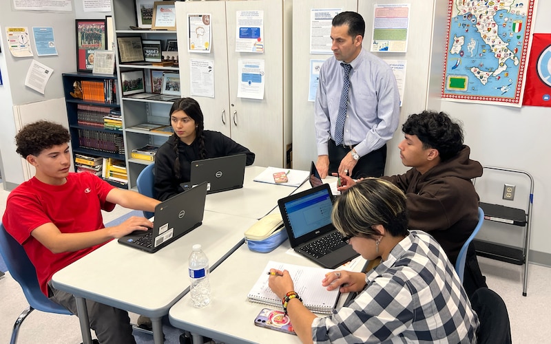 A man in a shirt and tie talks to four students at tables.