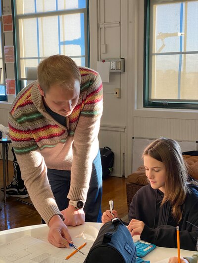An adult teacher stands while a student sits at a table working on classwork. There are windows and a white wall in the background.