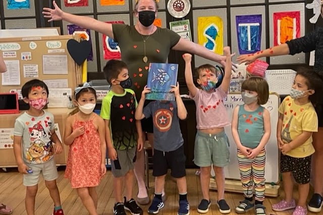 A woman wearing a mask stretches out her arms as several children stand below her while they all wear masks and stand in front of a wall with letters on it. 