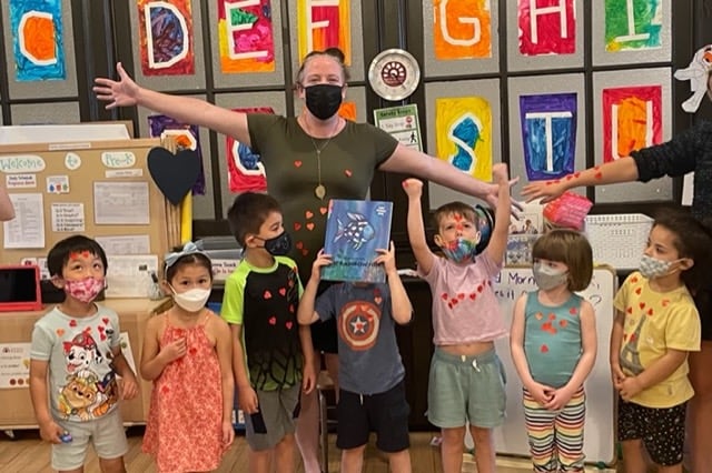 A woman wearing a mask stretches out her arms as several children stand below her while they all wear masks and stand in front of a wall with letters on it. 