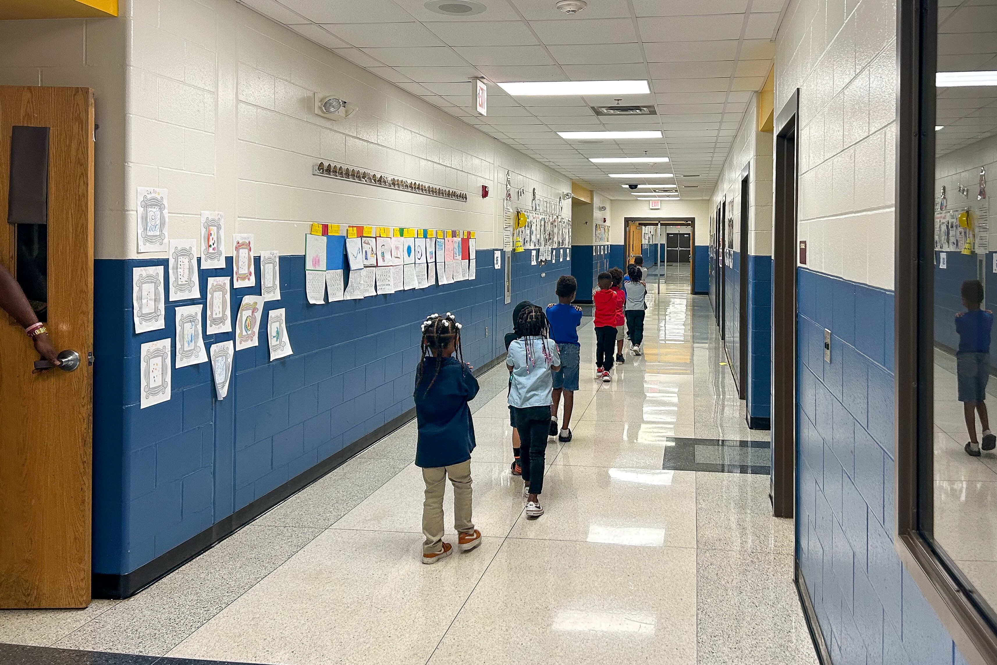 A line of young children walk down the hall of a school hallway.