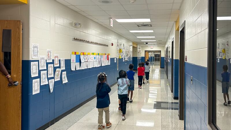 A line of young children walk down the hall of a school hallway.