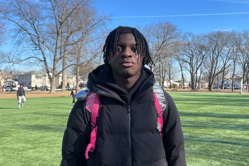 A Black high school student with short dark hair and wearing a black jacket poses for a portrait outside on a cold and sunny day.