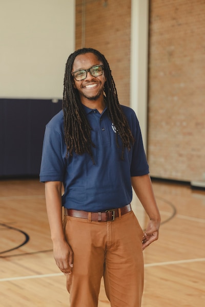 A Black man with long dark hair and wearing a blue dress shirt and tan dress pants smiles and poses for a photograph while standing in the middle of a school gym floor.