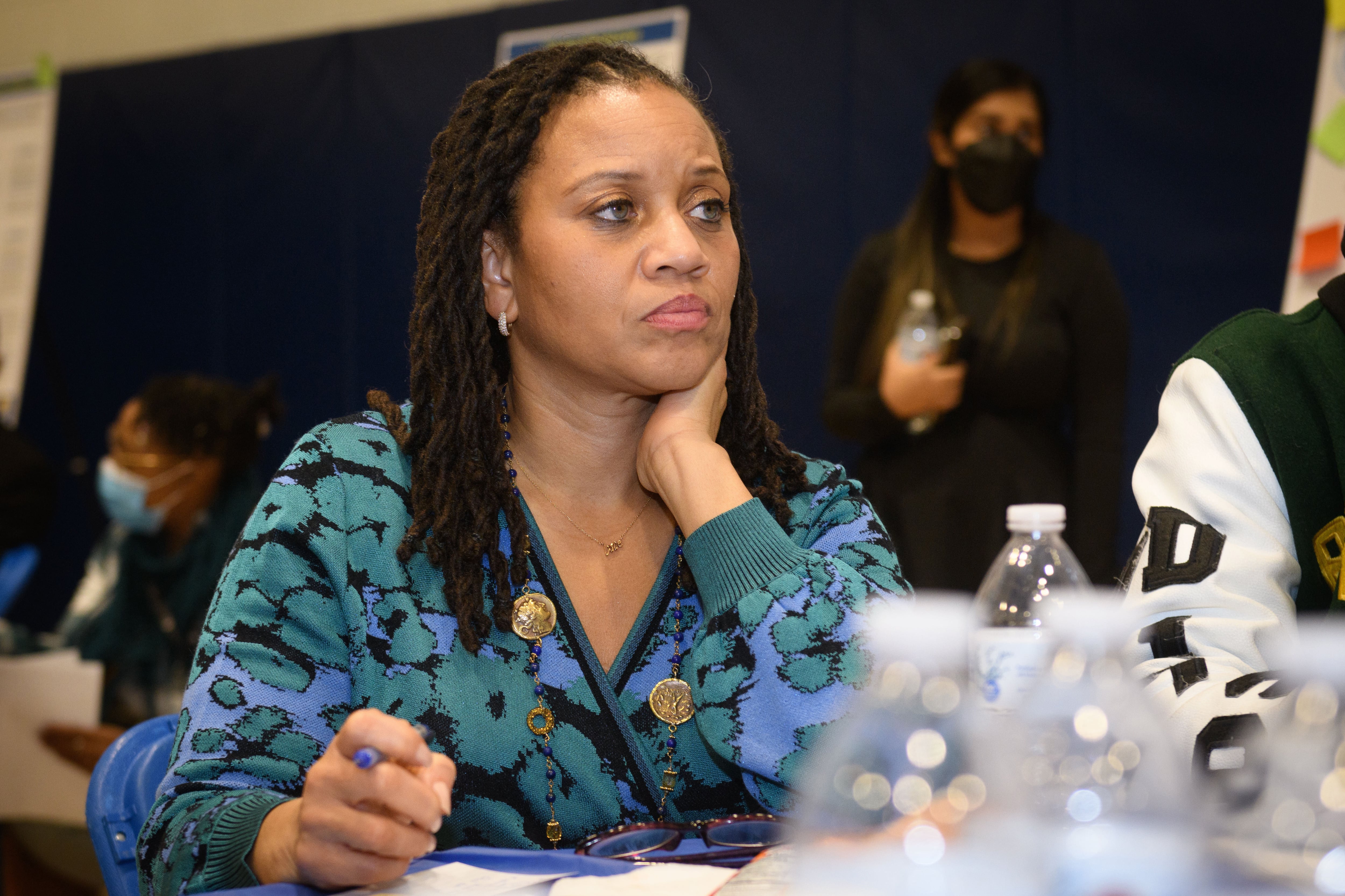 A woman with medium length dark hair and wearing a blue blouse sits at a table.