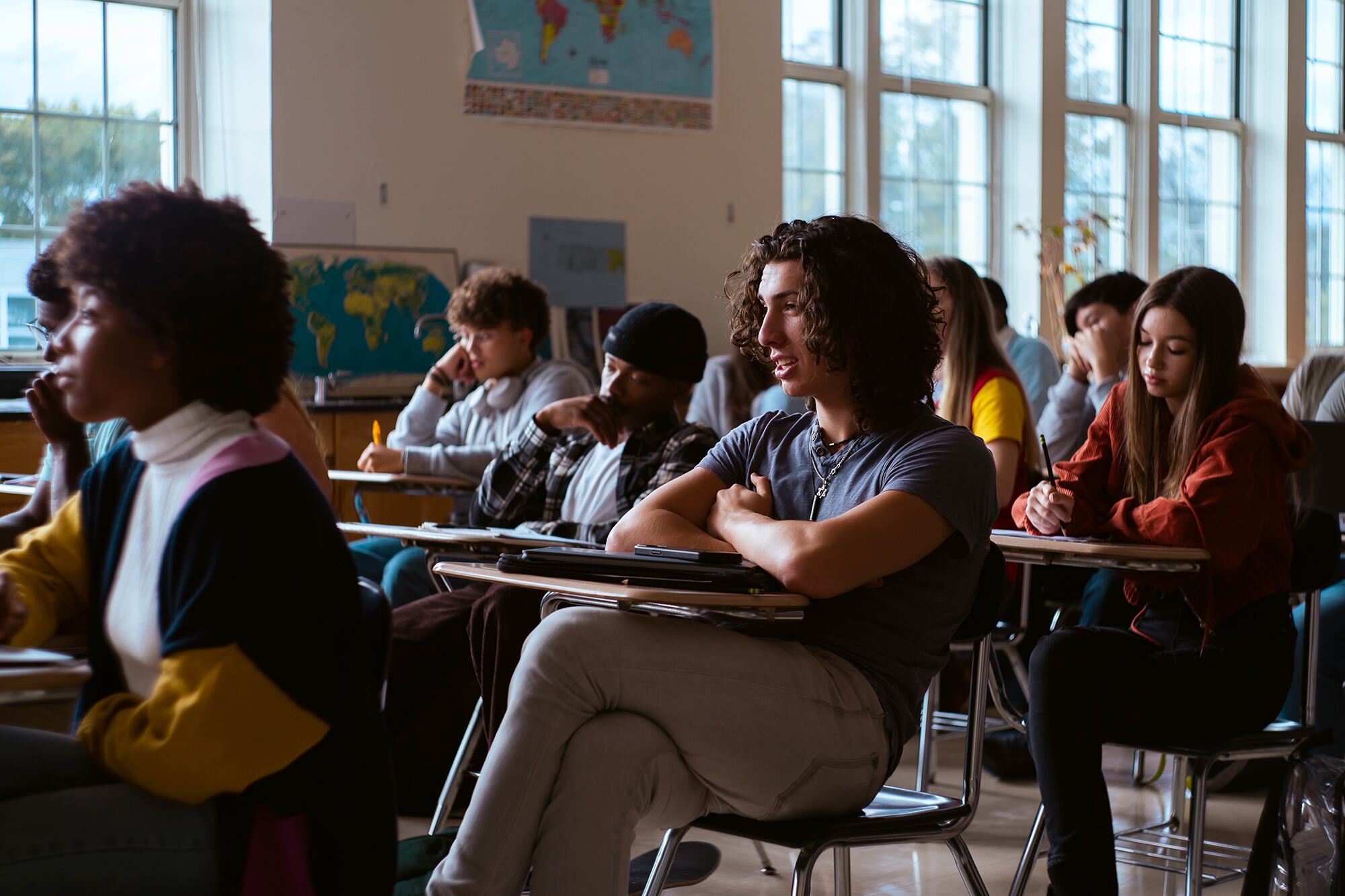Group of high school students sit at desks in classroom listening to teacher.