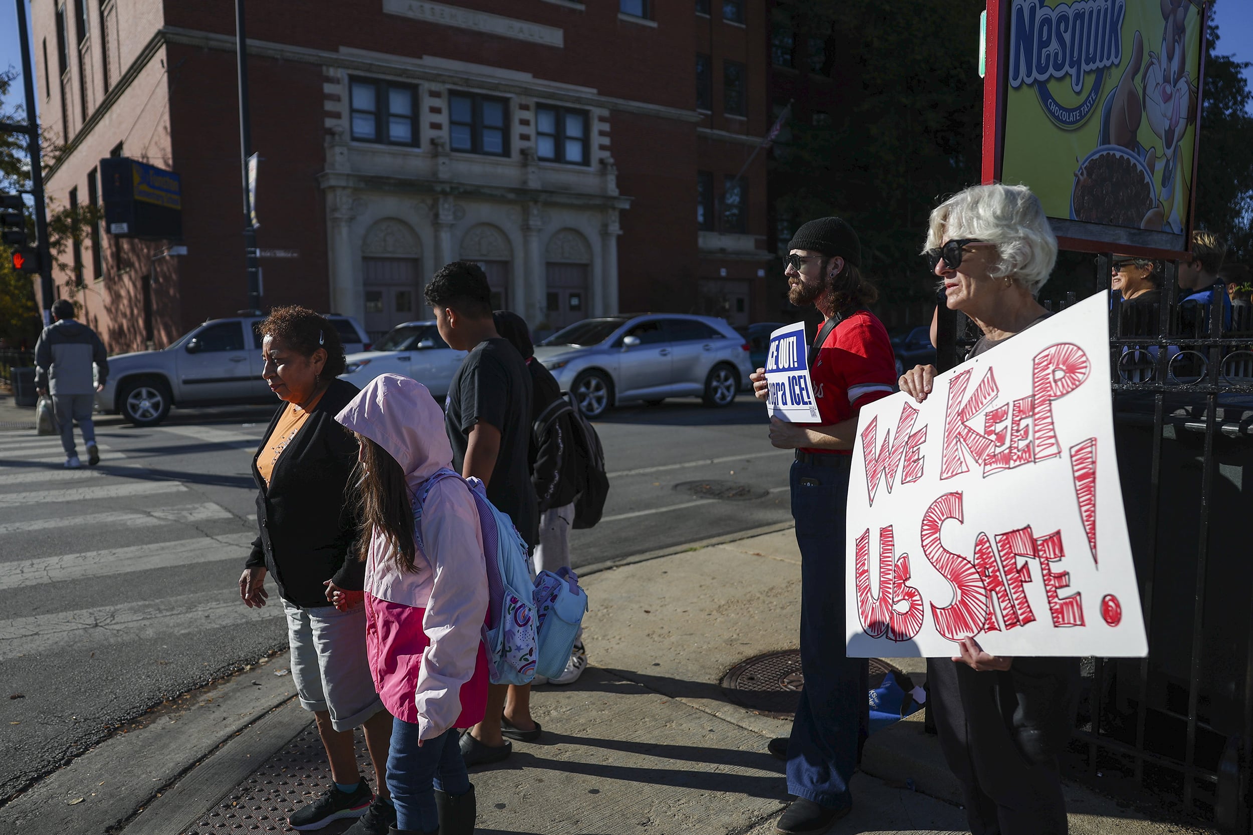 A photograph of two people holding up signs while a family waits at the crosswalk before crossing the street.