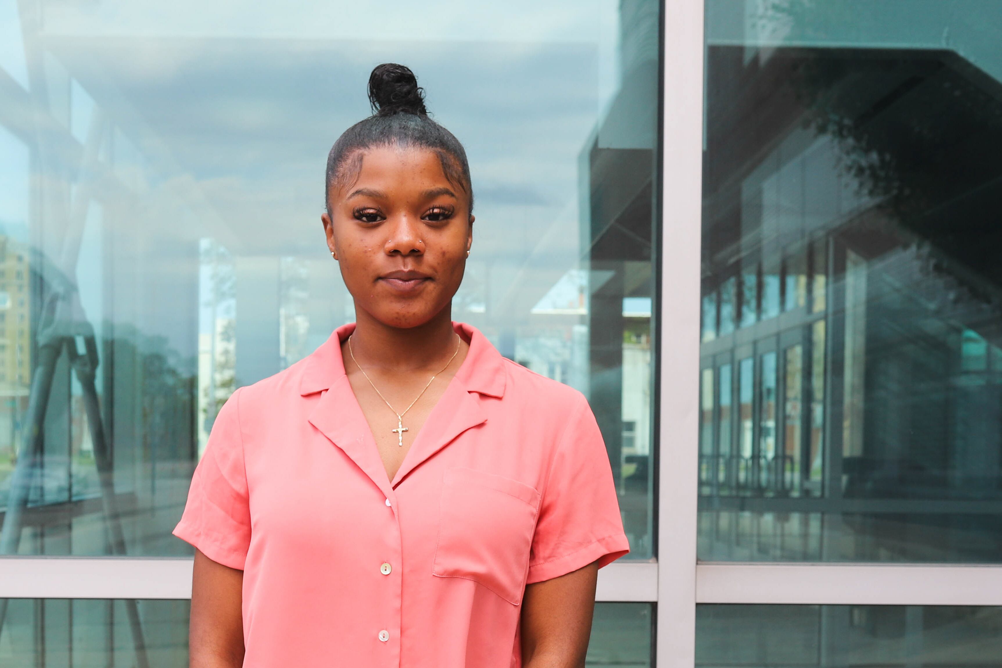 A young woman wearing a pink shirt and cross necklace stands in front of a building with large glass windows.