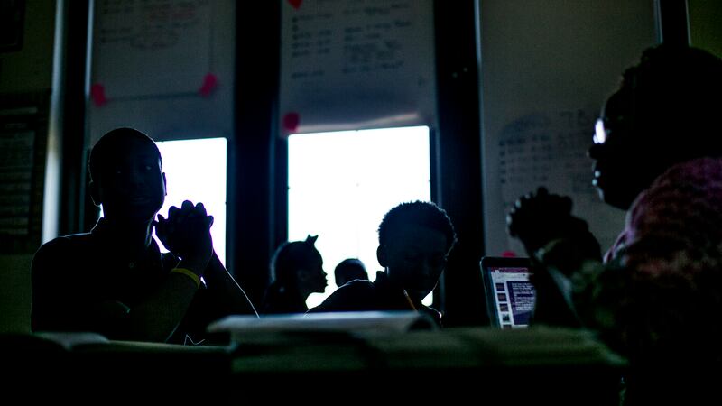 A silhouette of four students as they sit in chairs in a classroom in front of a bright window.