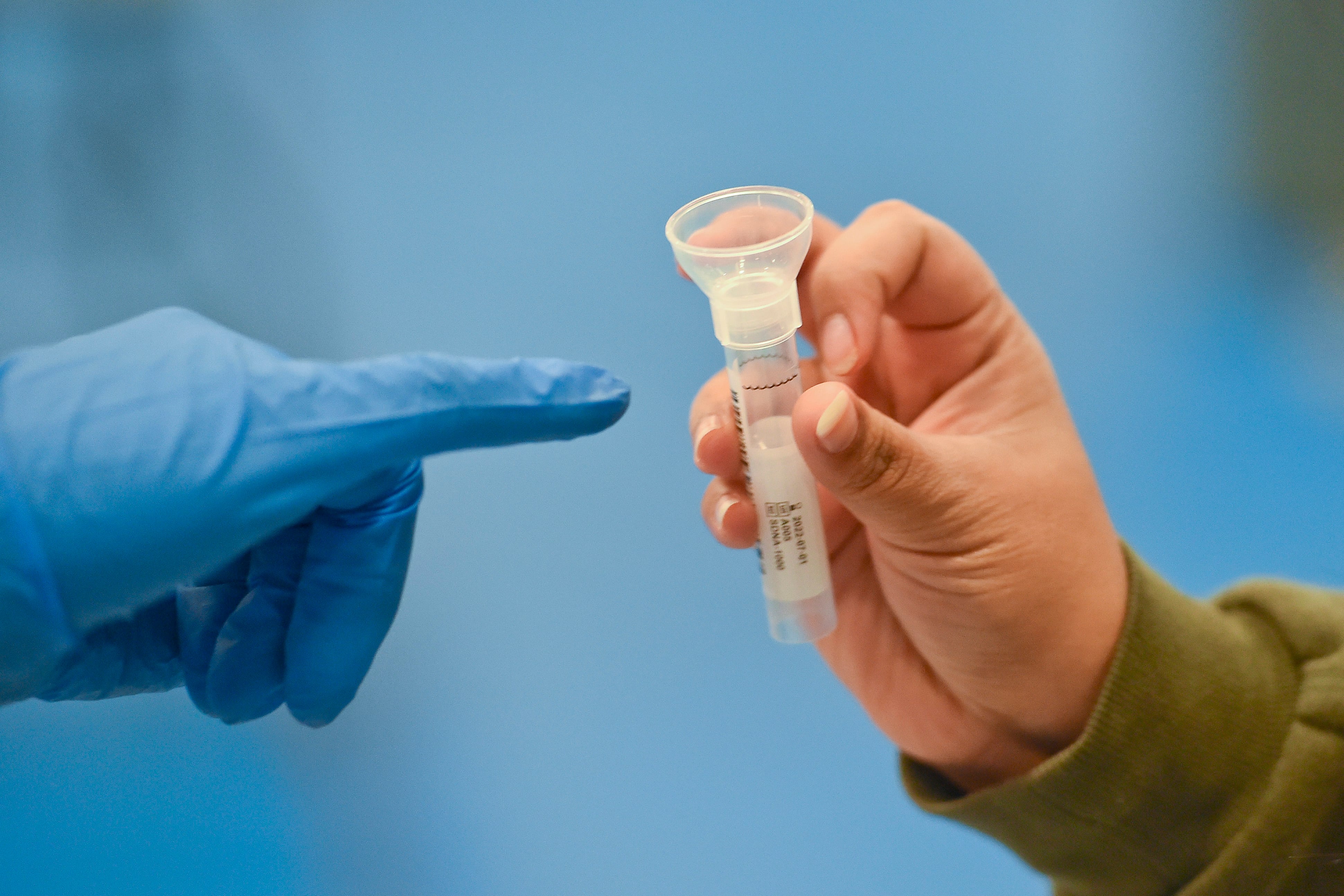 A hand with a blue latex glove points to a cylindrical saliva testing tube, used to detect COVID.