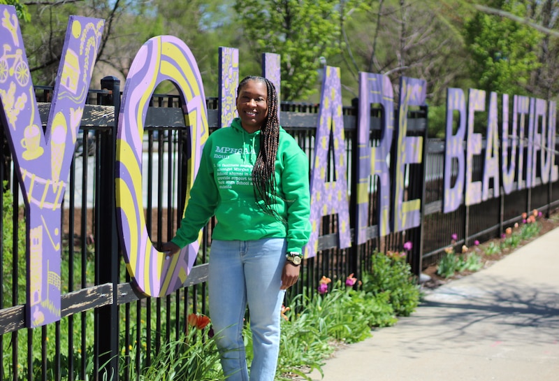 A woman with long dark hair and wearing a green hoodie, poses for a portrait in front a large sign outside.