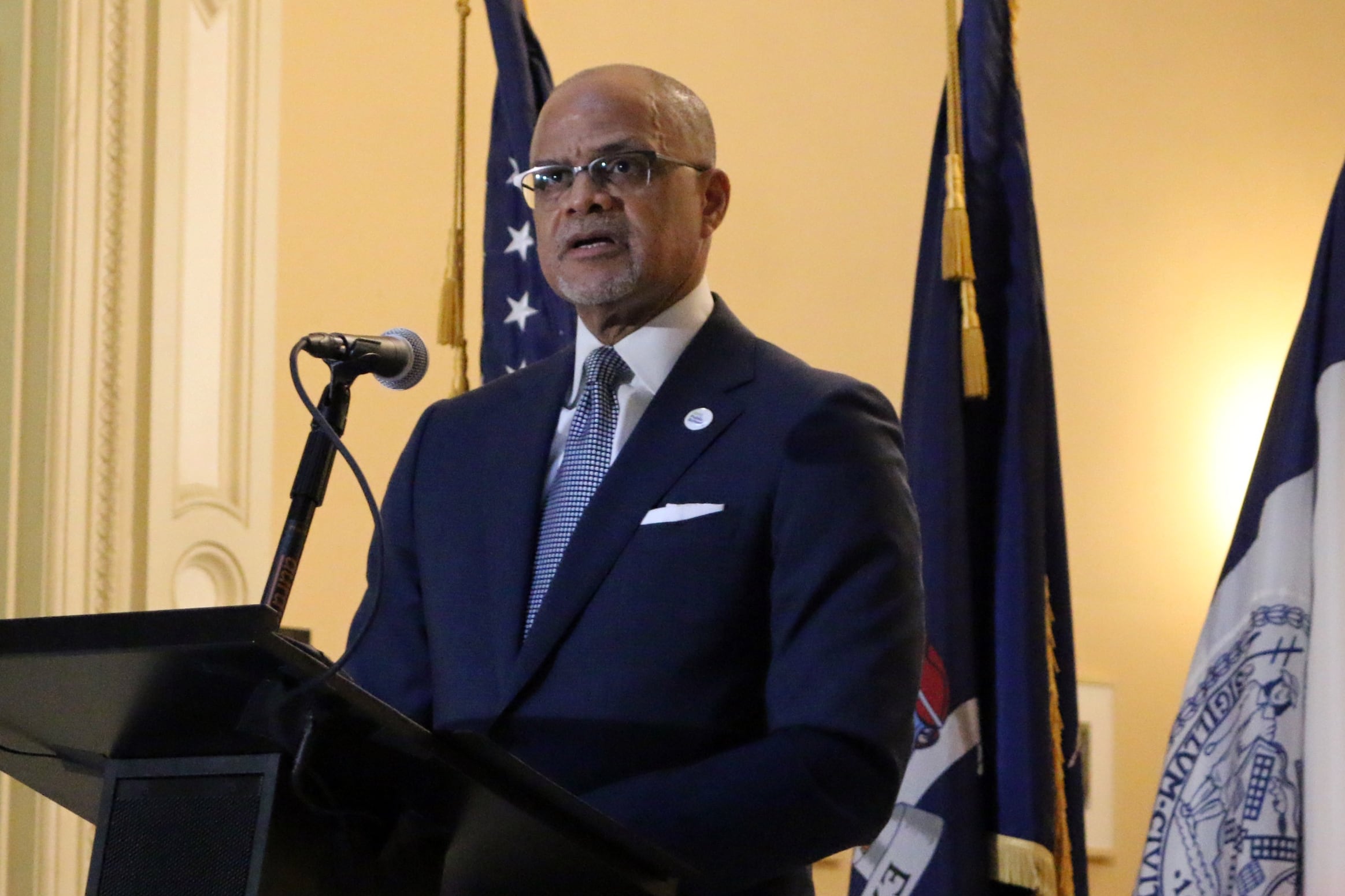 A man wearing glasses and a dark suit speaks from a podium with three flags in the background.