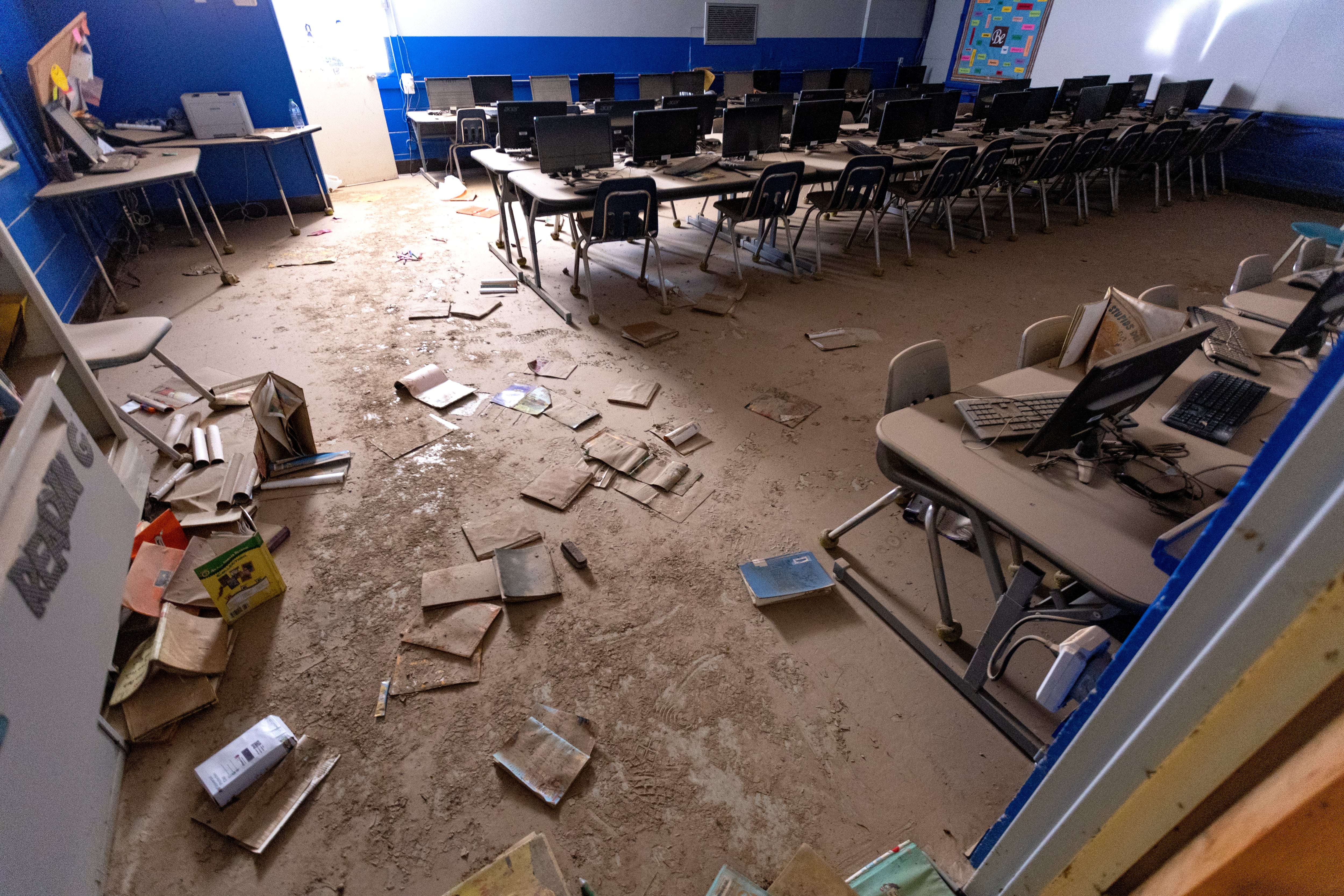 Mud-covered desks and computers and books on the floor inside of a elementary school classroom