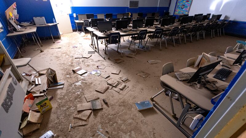 Mud-covered desks and computers and books on the floor inside of a elementary school classroom