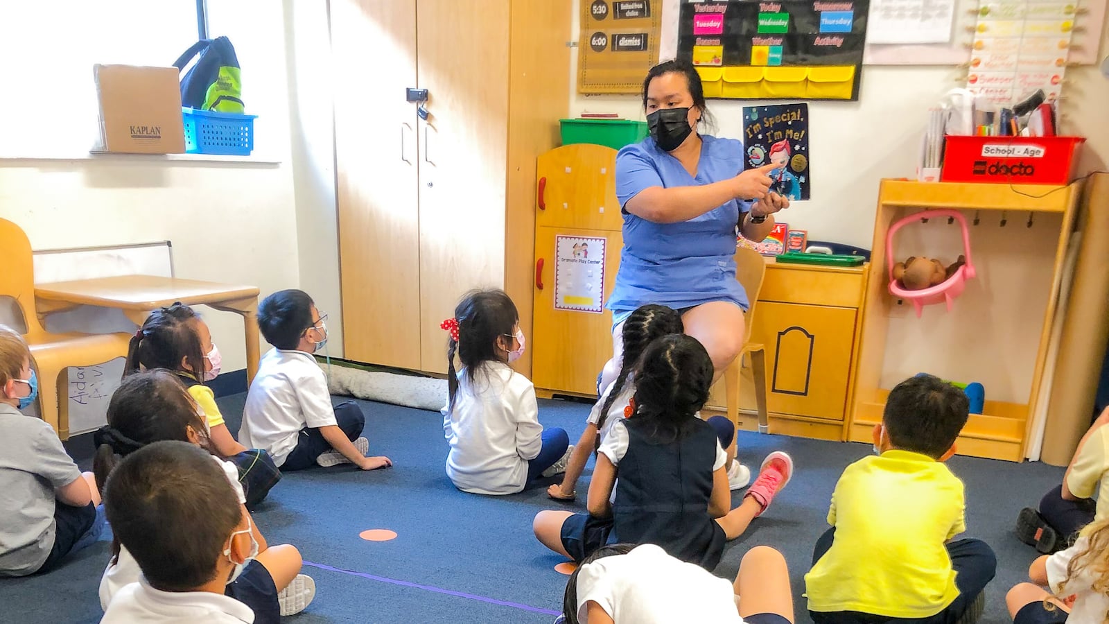 A masked teacher reads a book to students, who are seated on a rug in front of them.