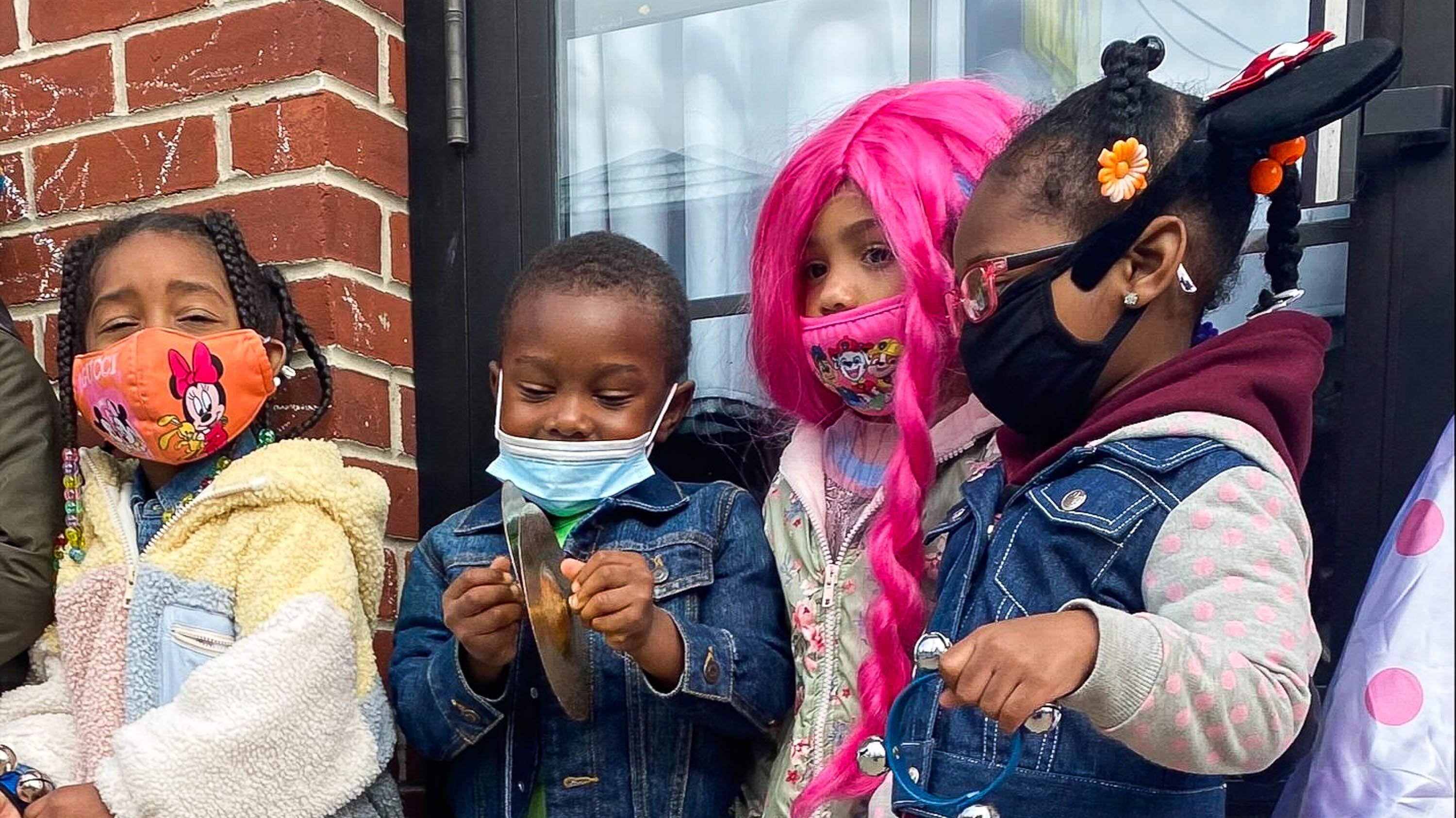 Four preschoolers, each wearing protective masks, stand side-by-side next to a brick wall and window.