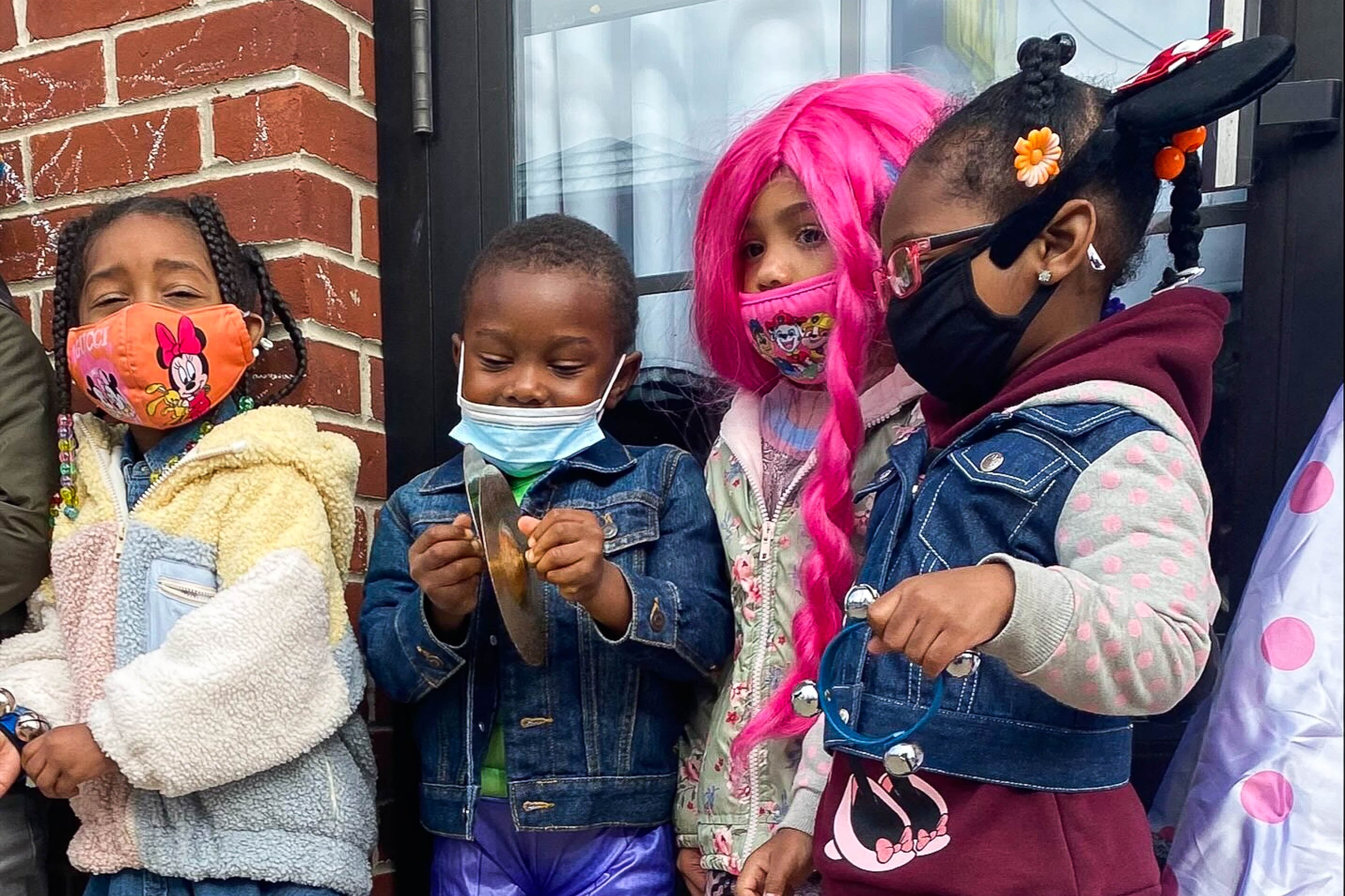 Four preschoolers, each wearing protective masks, stand side-by-side next to a brick wall and window.