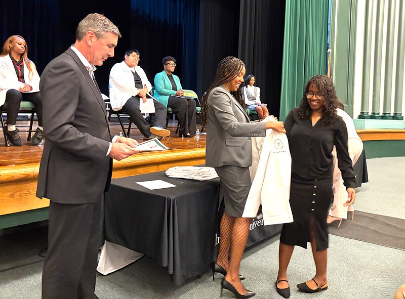 A young woman in dark dress is handed a white lab coat.