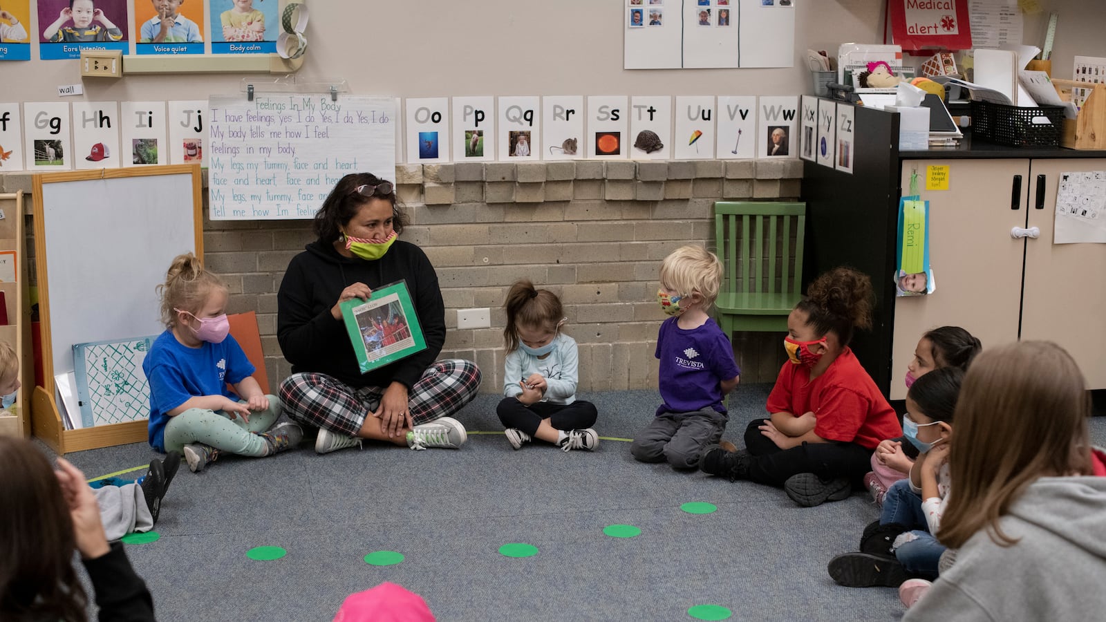 A preschool teacher sits with her students on a carpet lined with green dots, holding a picture as she speaks with the children.