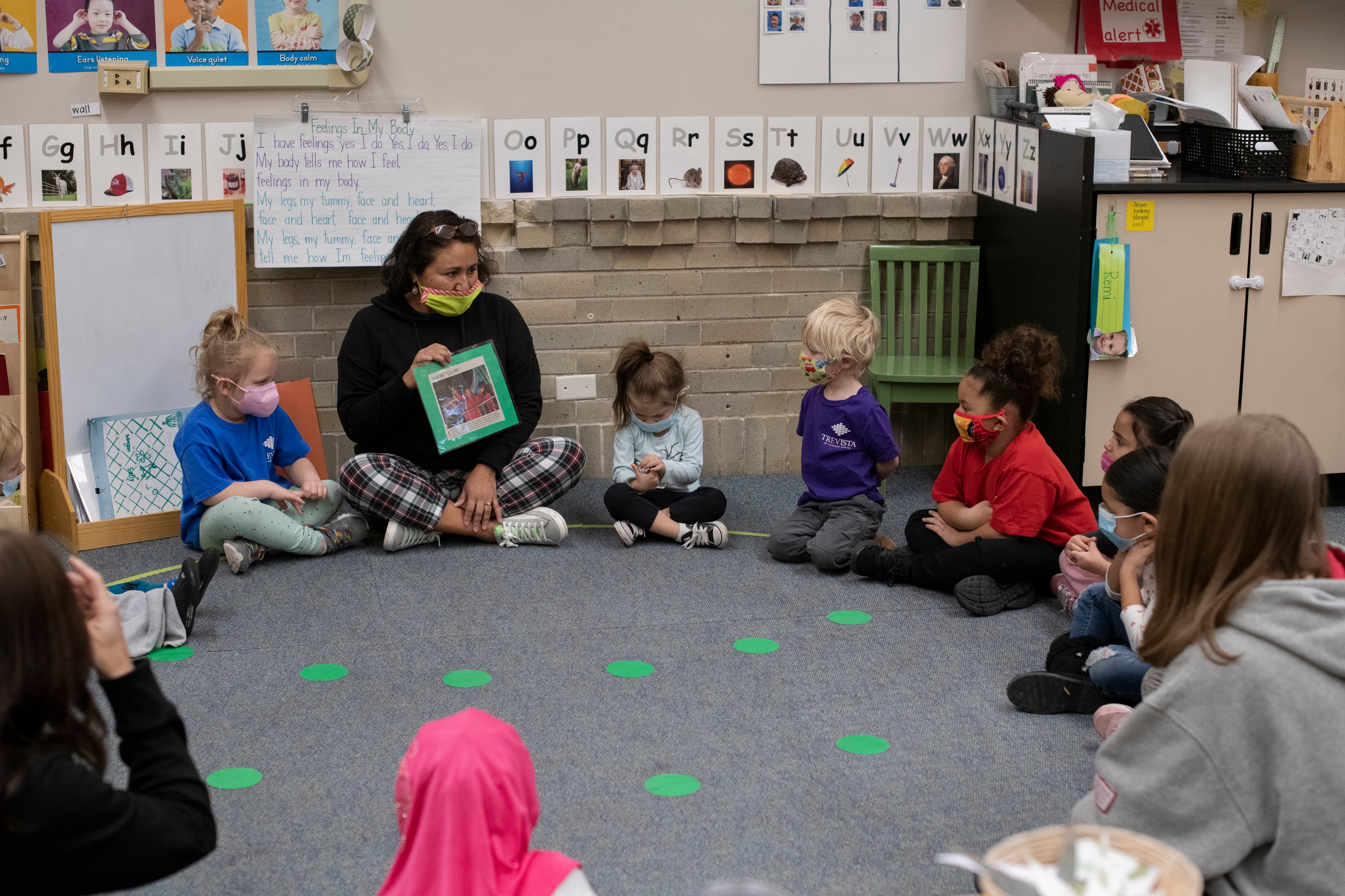 A preschool teacher sits with her students on a carpet lined with green dots, holding a picture as she speaks with the children.