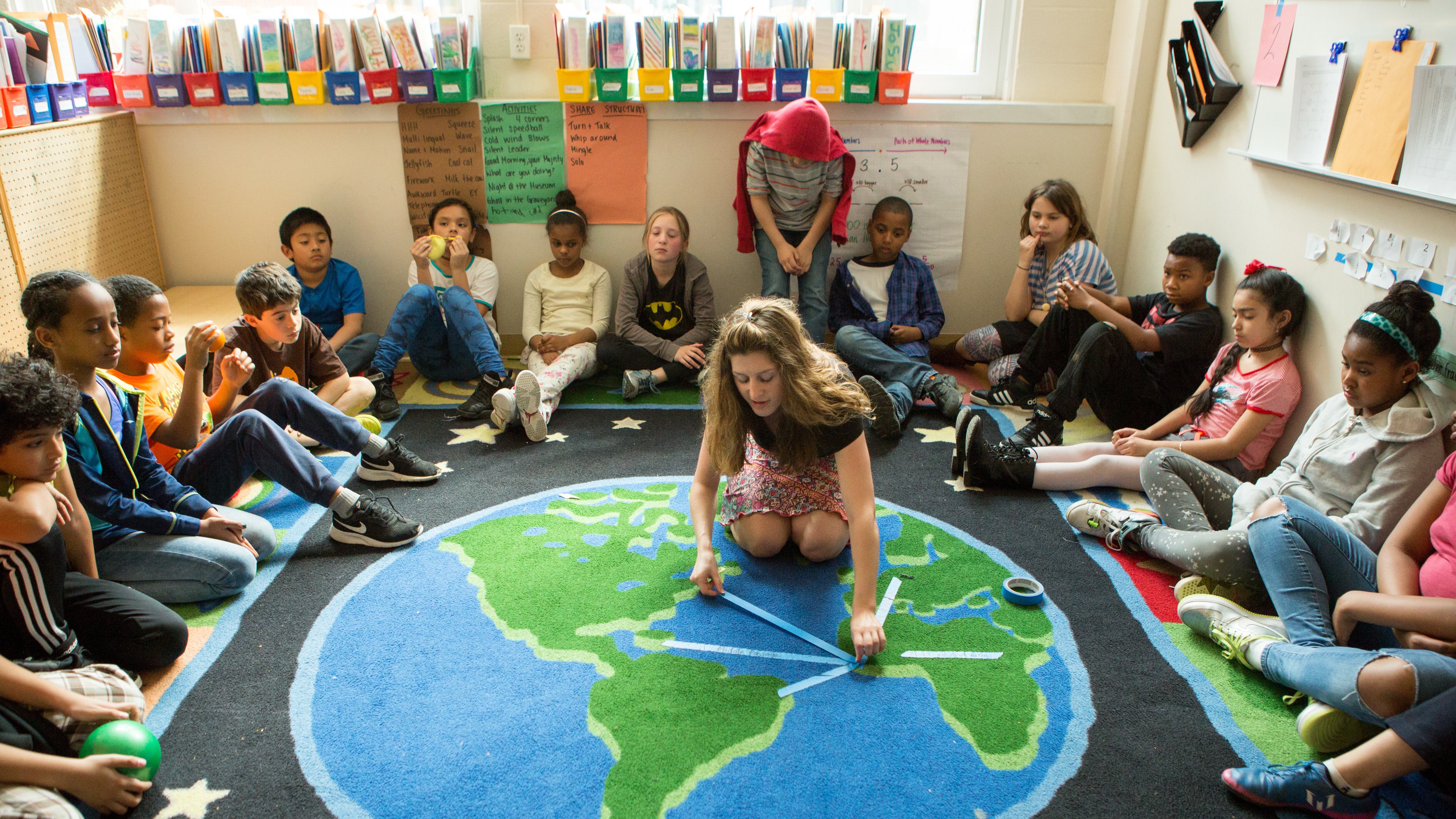 A teacher kneels on a rug shaped like the globe while students sit in a circle around her.