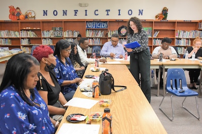 Men and women review documents from their seats at a table while a woman with curly hair and glasses reads from a blue folder she is holding.